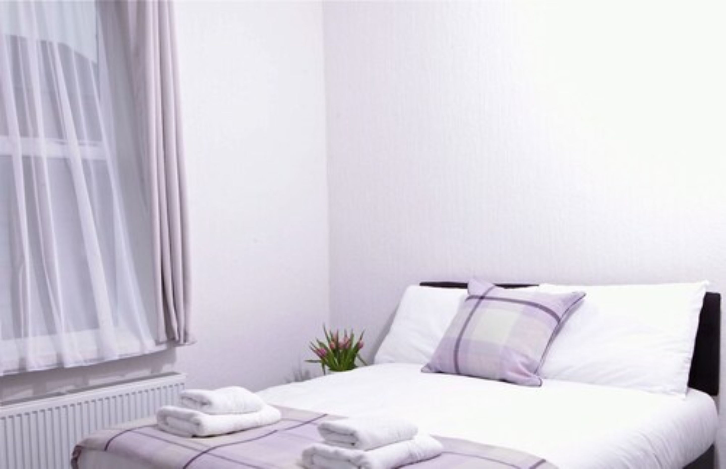 A neatly made bed with white and purple bedding, a small bedside plant, and a window with sheer curtains.
