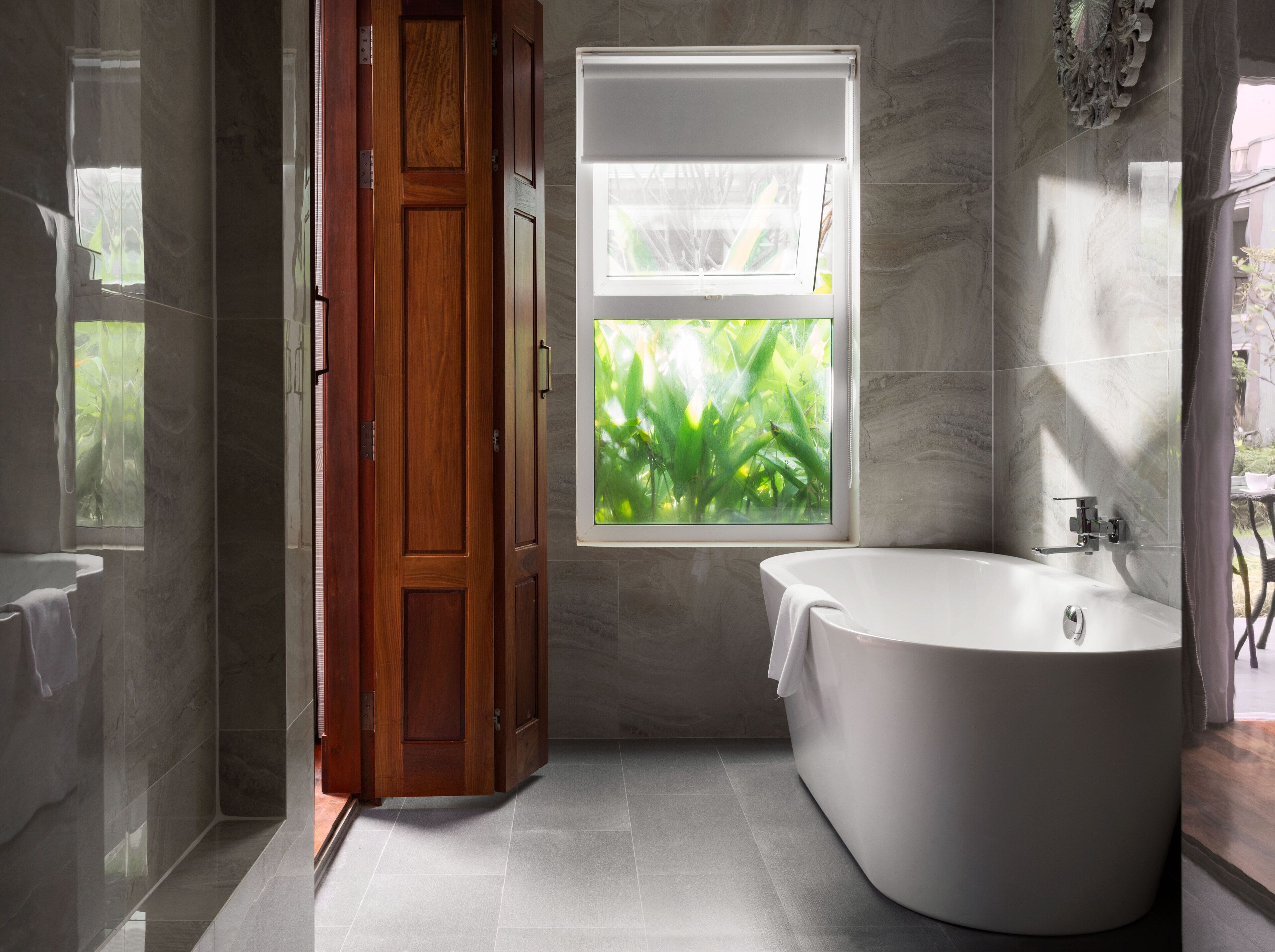 A modern bathroom with a freestanding bathtub, a wooden door, and a window with a view of greenery.