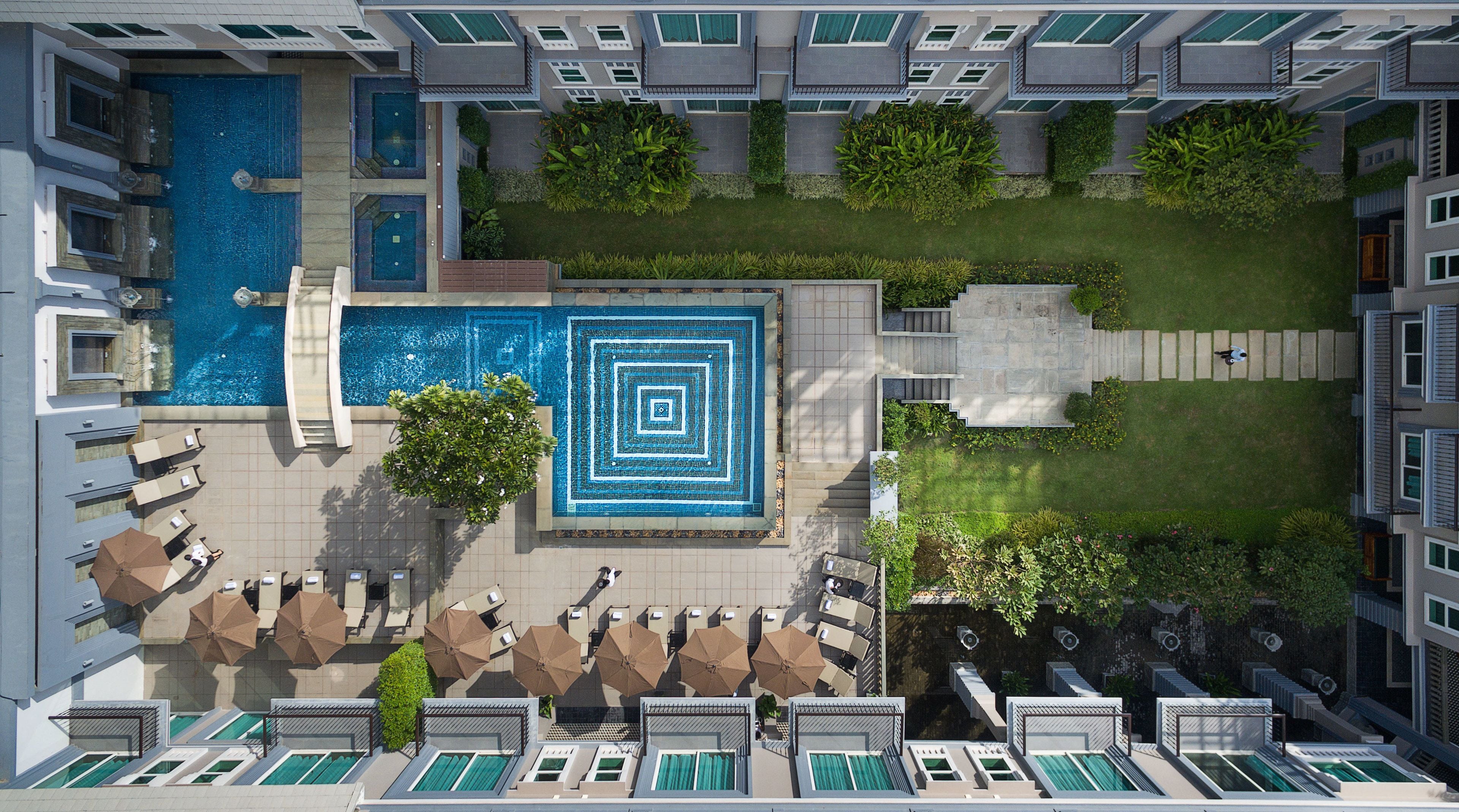 An aerial view of a hotel with a central swimming pool, multiple lounge areas with umbrellas, and a well-maintained garden.