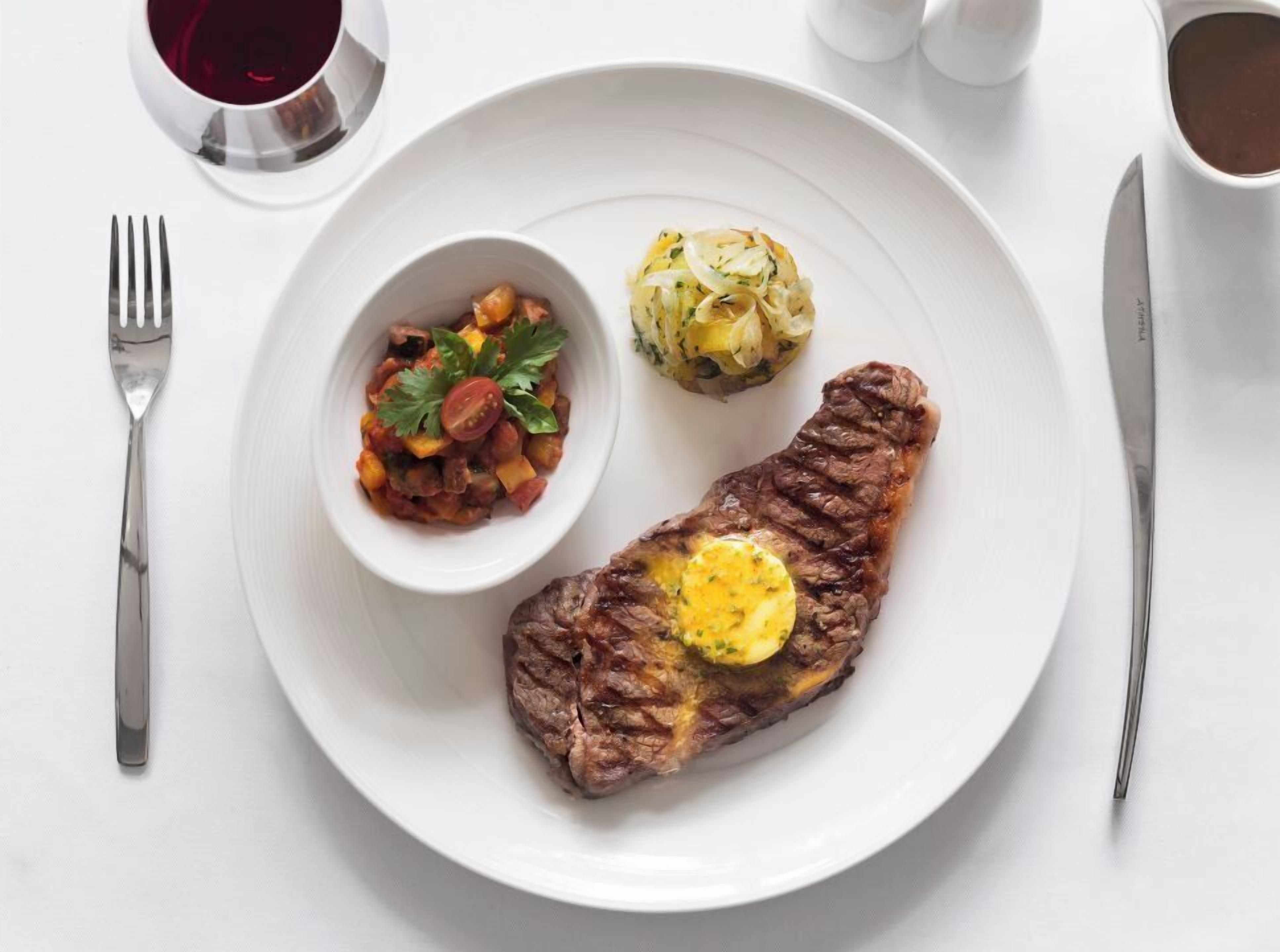 A plate with a steak topped with butter, a side of vegetables, and a small bowl of sauce.