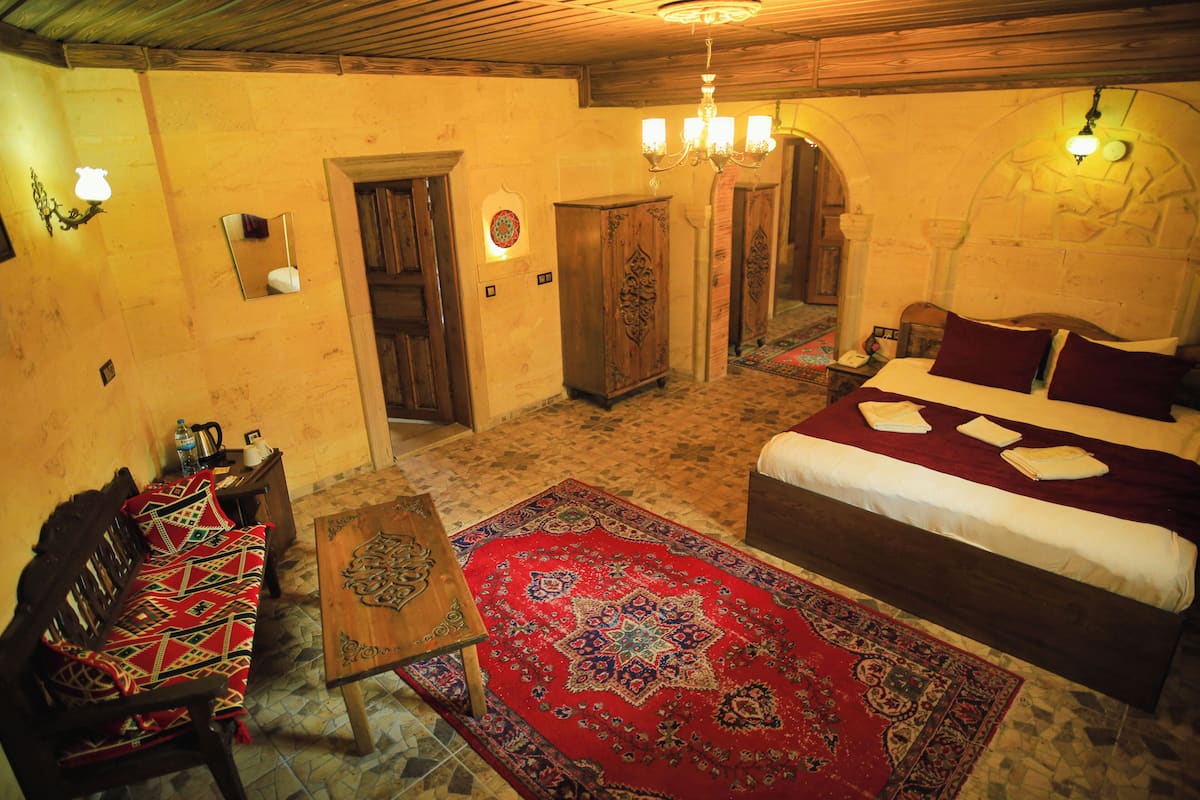Una habitación tradicional con una cama, un banco de madera, un tapiz rojo estampado y una araña.