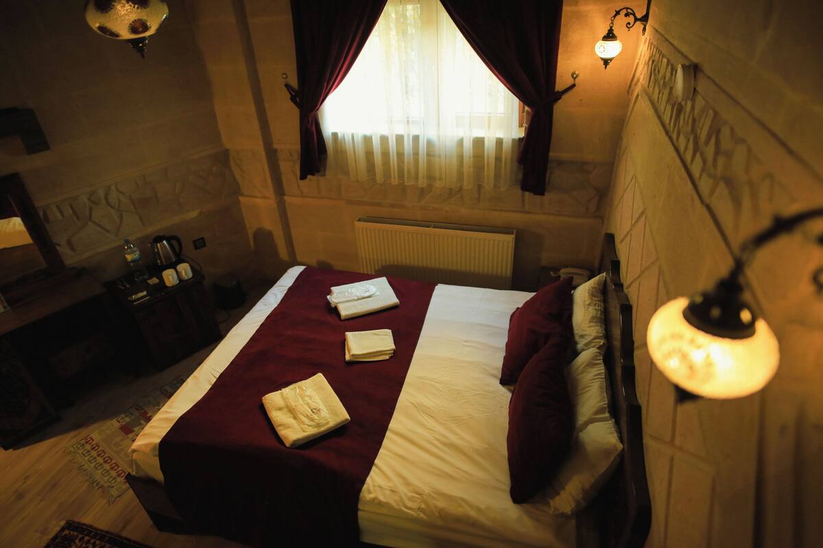 Habitación de hotel con cama, mesita de noche, lámpara, radiador y ventana con cortinas.