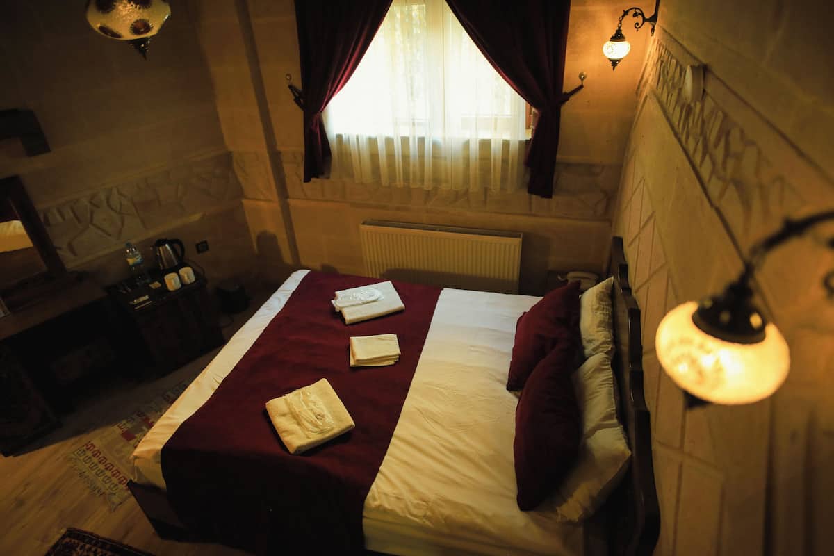 Habitación de hotel con cama, mesita de noche, lámpara, radiador y ventana con cortinas.