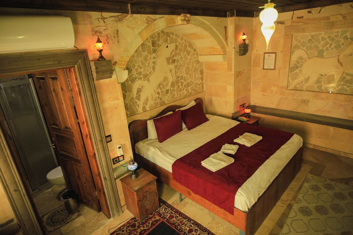 Un dormitorio con pared de piedra, una cama con colcha roja, cabecero de madera y una mesita de noche con lámpara.
