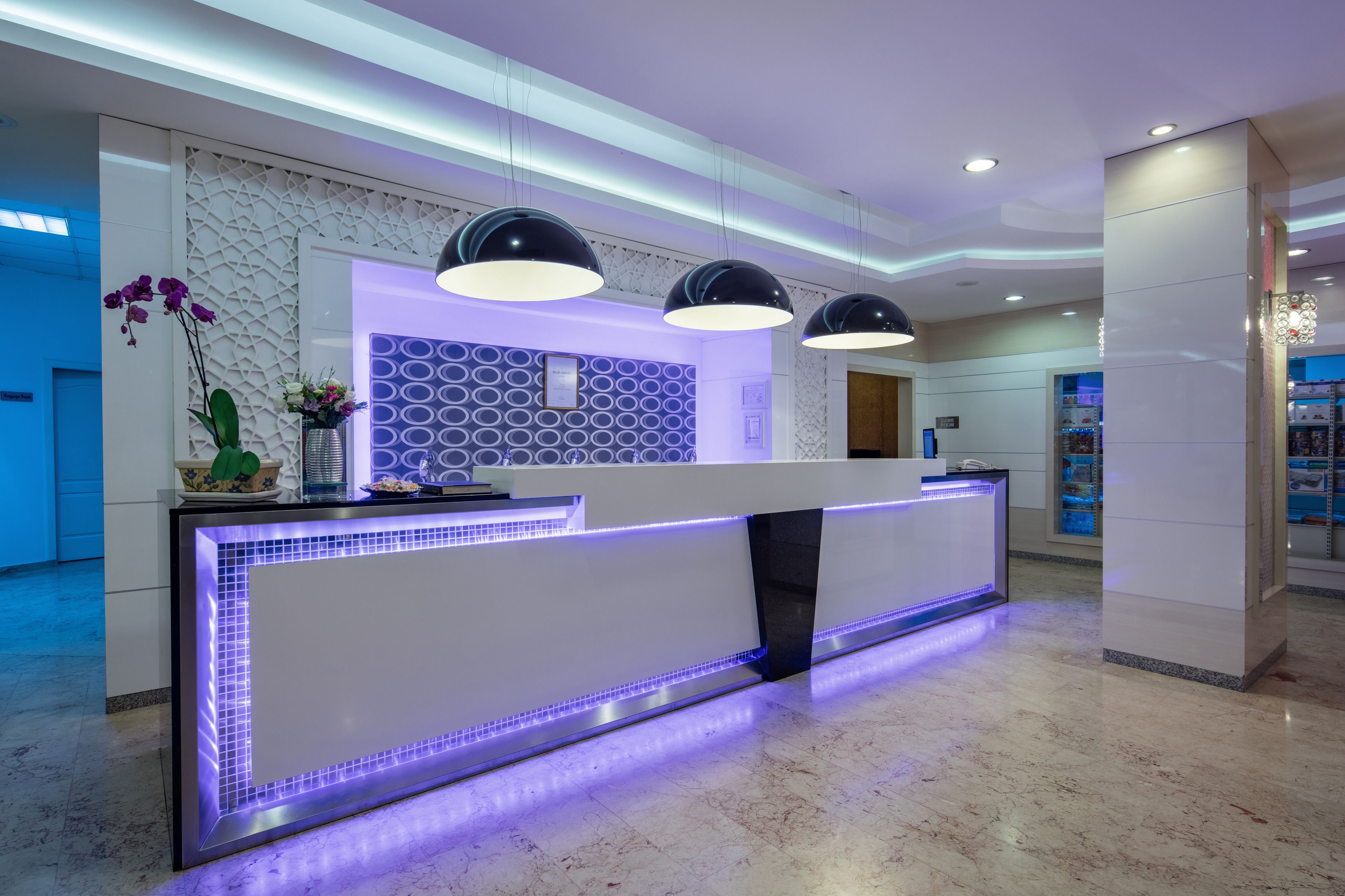 A modern reception desk with a white counter and blue LED lighting, surrounded by a decorative wall with circular patterns and a large potted plant.