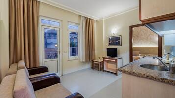 A hotel room with a sofa, a TV, a small table, a kitchen area with a sink, and a balcony view.