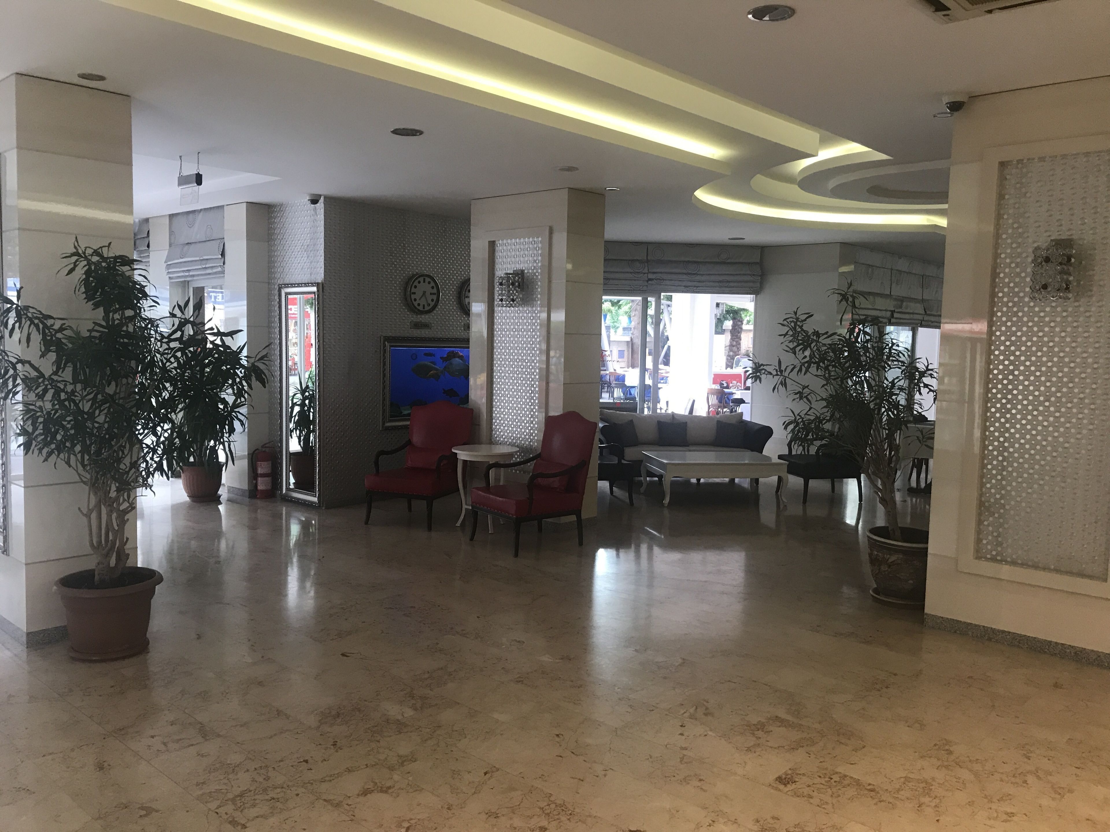 A spacious lobby with a modern design, featuring a seating area with red chairs and a white sofa, a large wall-mounted clock, and a potted plant.