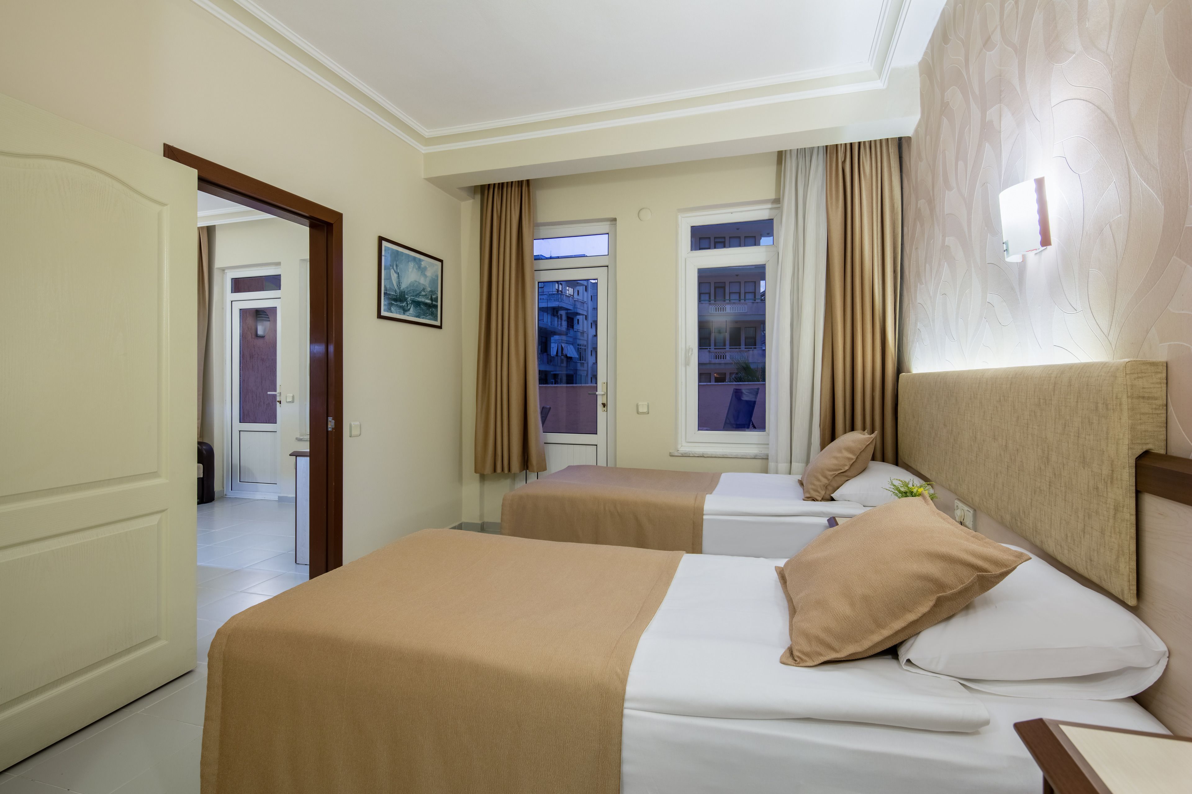 A hotel room with two beds, a large window, a door leading to another room, and a wall-mounted light fixture.