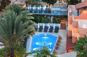 Una piscina en la azotea con sillones de descanso y sombrillas, rodeada de palmeras y un muro de piedra.