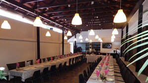 Una larga mesa de comedor dispuesta con manteles blancos y servilletas rosas, rodeada de sillas, en una habitación con suelo de piedra y lámparas colgantes.