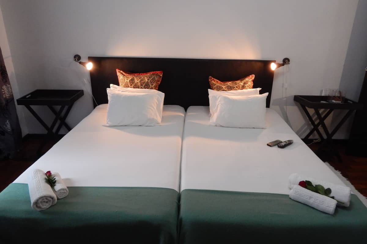 Una cama doble con sábanas blancas, dos almohadones decorativos, una mesita de noche y una colcha verde.