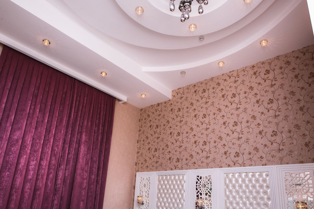 Un dormitorio lujoso con una cama blanca, dos sillas blancas, un techo decorativo y un muro estampado.