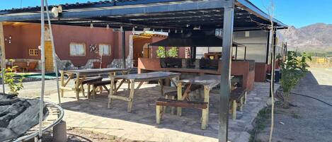 Un patio al aire libre con mesas y bancos de madera rústica, una parrilla y una zona de estar cubierta con vistas a las montañas al fondo.