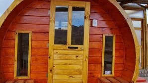 Une cabane en bois à la forme circulaire, dotée d’une porte centrale et de deux fenêtres de chaque côté, ainsi que d’un petit banc de chaque côté de la porte.