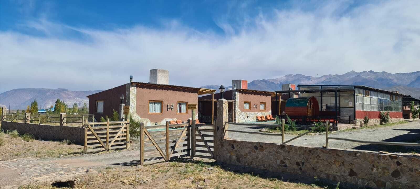 Un edificio rústico con pared de piedra y puerta de madera, una construcción pequeña con chimenea, y un edificio moderno de vidrio con un letrero.