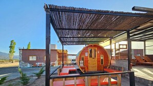 Une cabane rustique avec une façade en bois, une structure circulaire également en bois dotée d’une porte, et un espace de repos couvert avec des bancs.