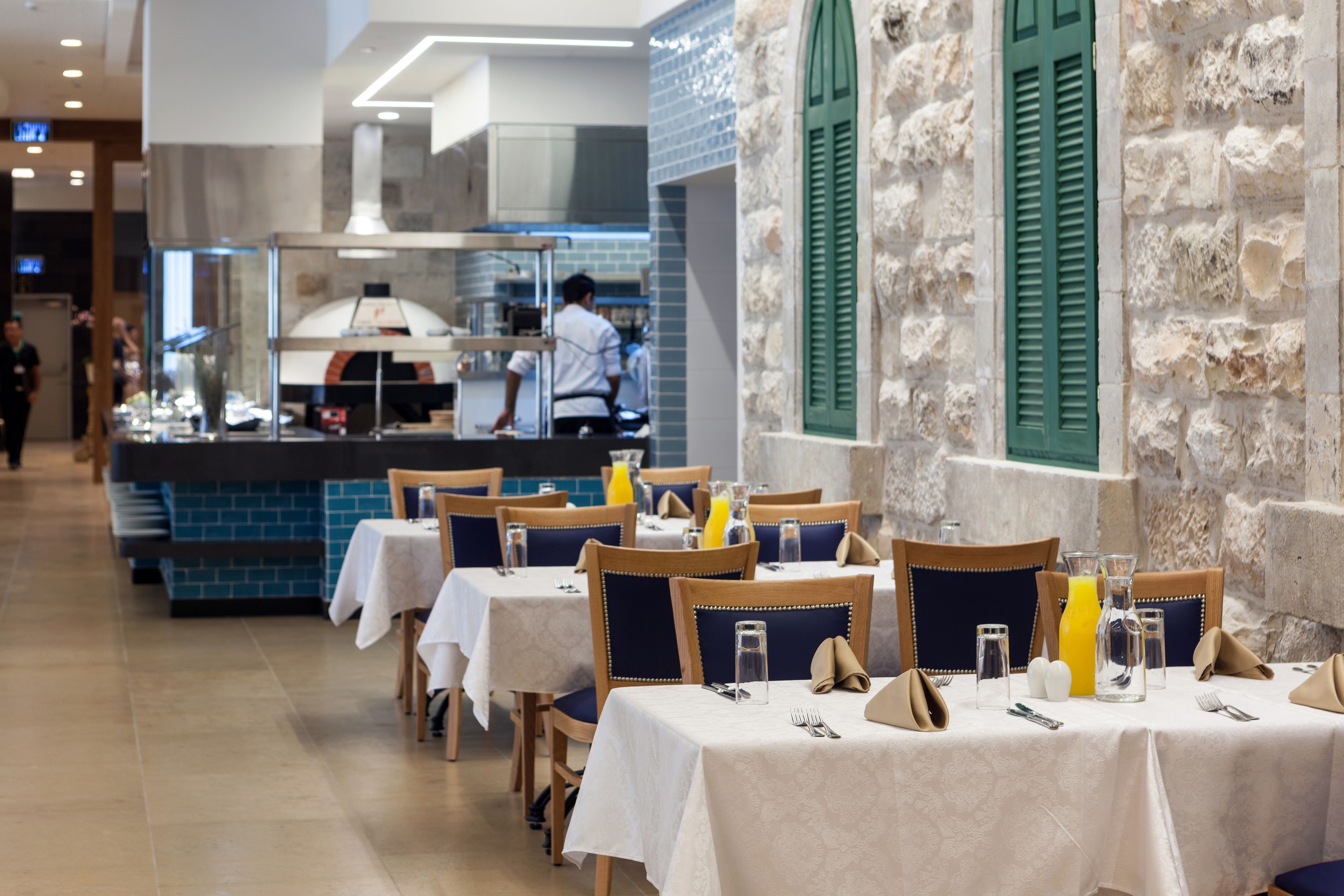 Un restaurant avec des tables dressées pour le service, un espace cuisine et des murs en pierre agrémentés de volets verts.