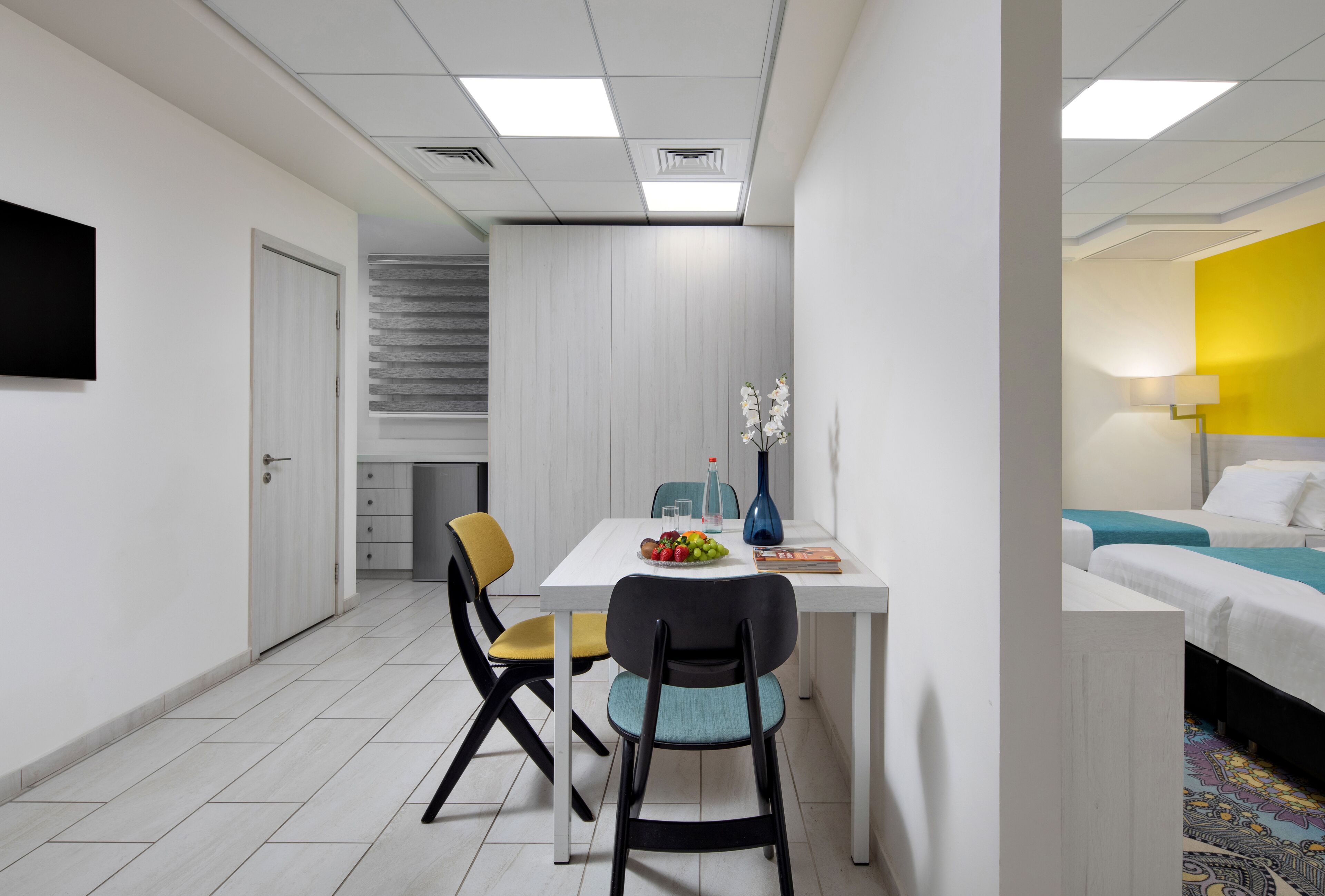 Una habitación de hotel moderna con una cama blanca, un escritorio pequeño con un florero y un área de comedor con sillas y una mesa.