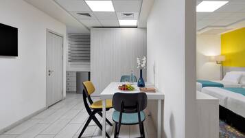 Una habitación de hotel moderna con una cama blanca, un escritorio pequeño con un florero y un área de comedor con sillas y una mesa.