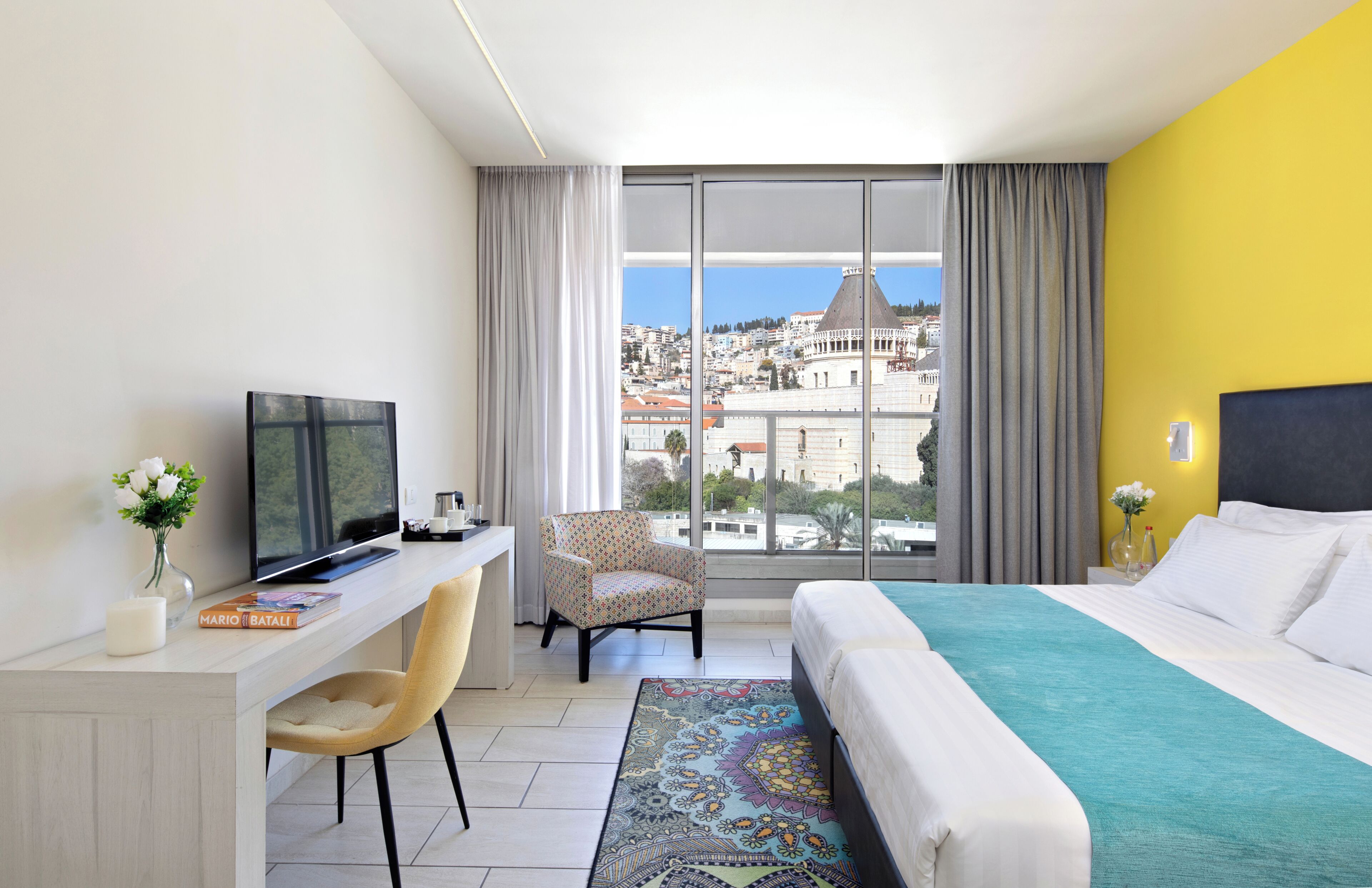A hotel room with a large window, a bed, a desk with a TV, a chair, and a view of the city.