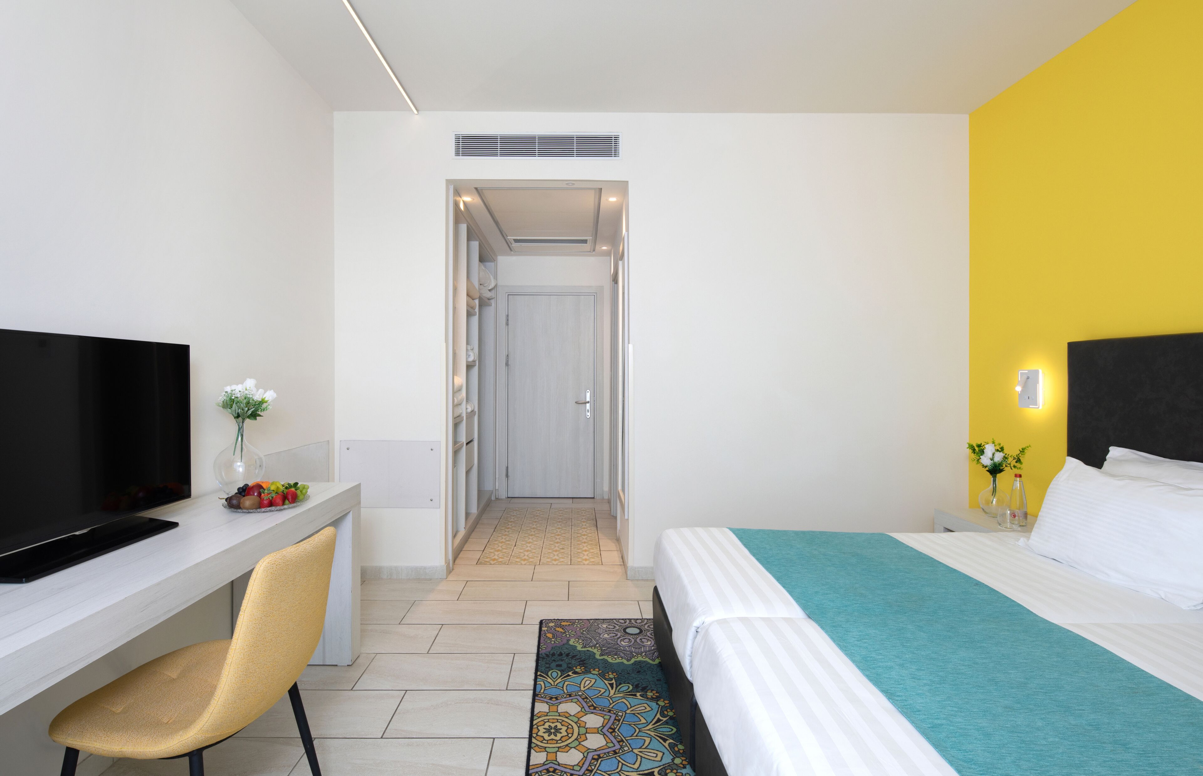 A hotel room with a bed, a desk with a TV, a chair, and a small table with flowers.