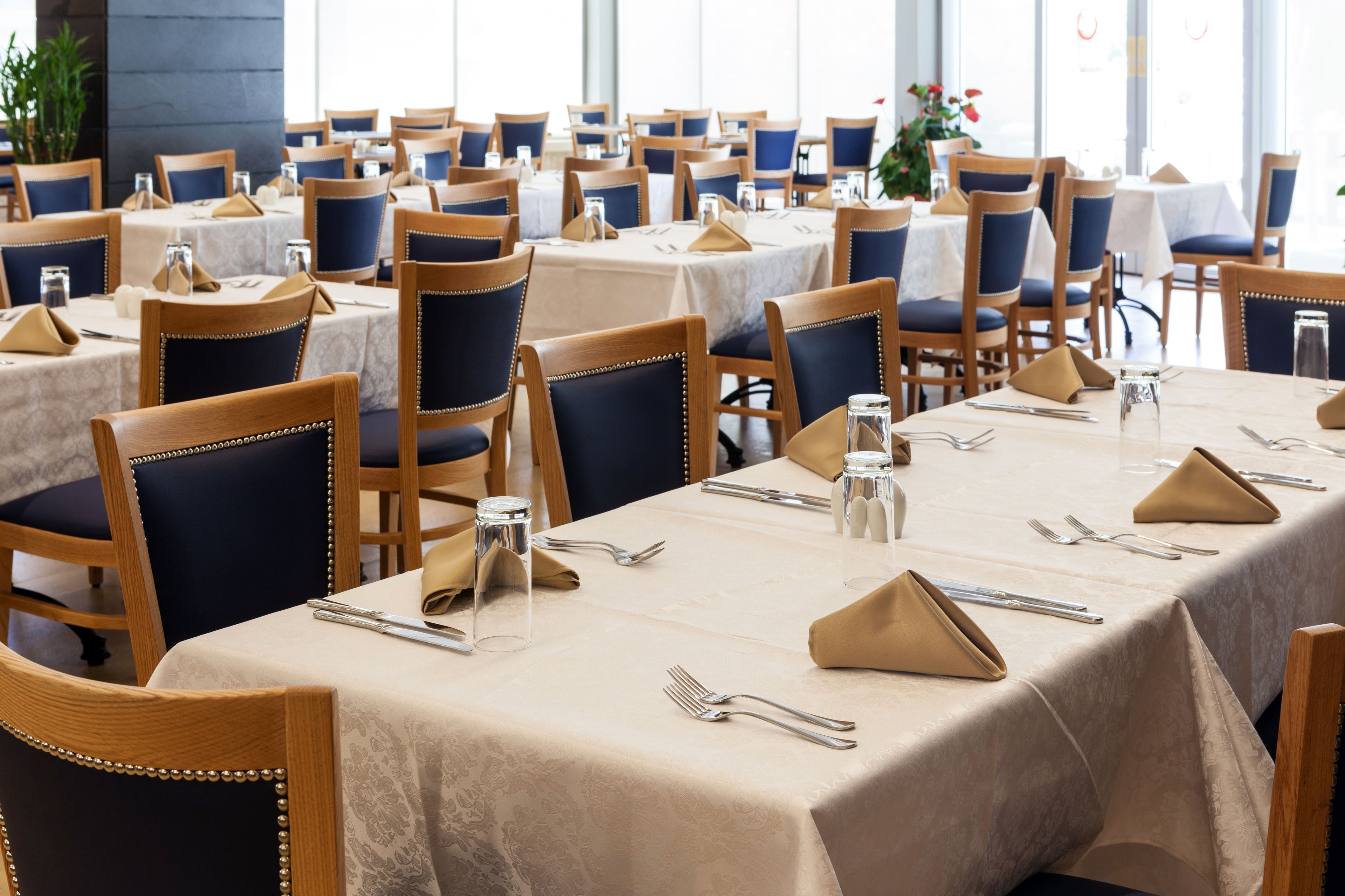A restaurant with multiple tables, each set with beige tablecloths, wooden chairs, and silverware.