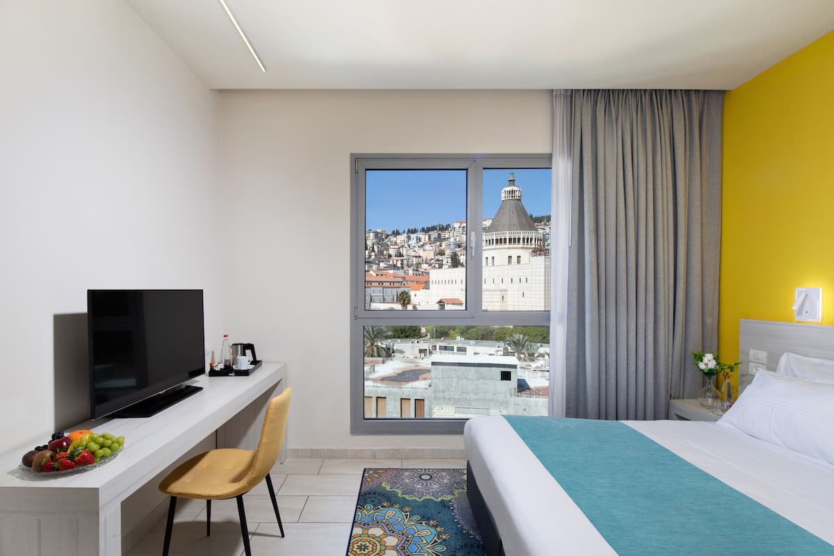 Ein Hotelzimmer mit einem Bett, einem Schreibtisch mit Fernseher, einem Sessel, einem Fenster mit Blick auf die Stadt und einem Vorhang.
