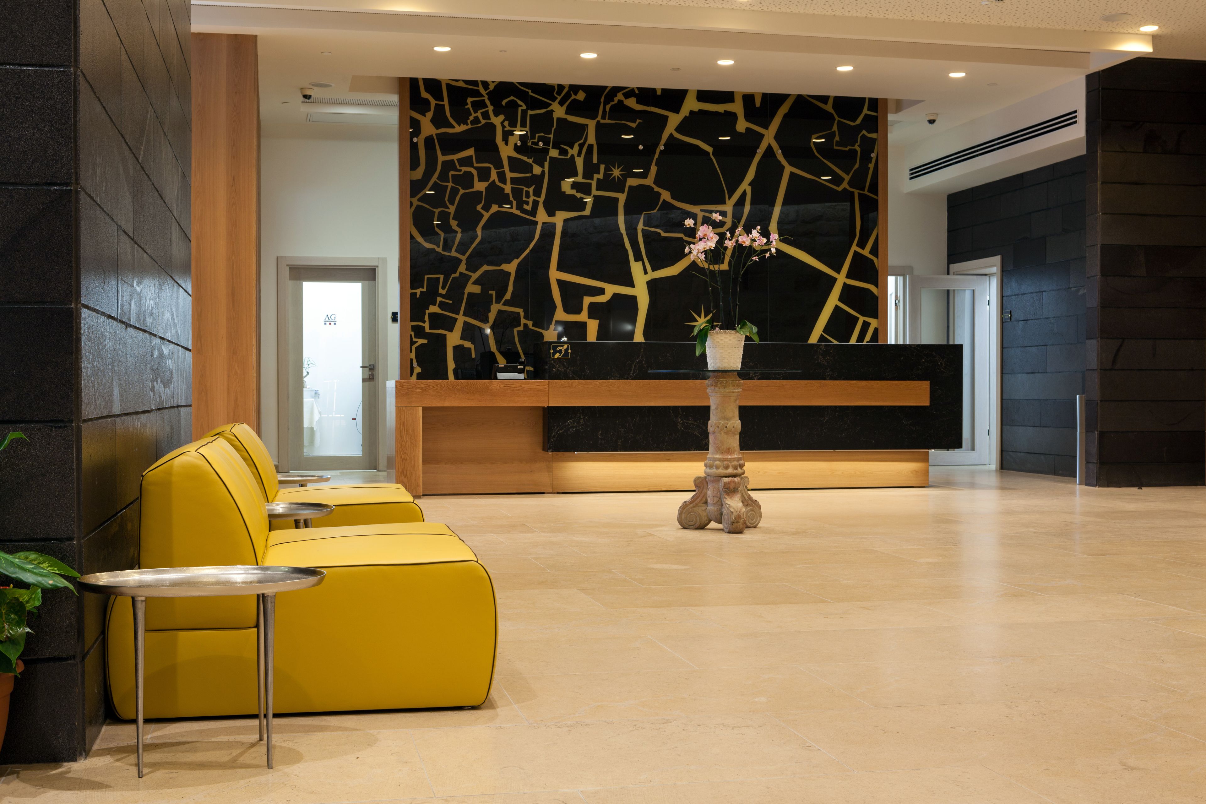 A reception area with a modern design, featuring a yellow sofa, a small round table, and a decorative pillar with a vase of flowers.