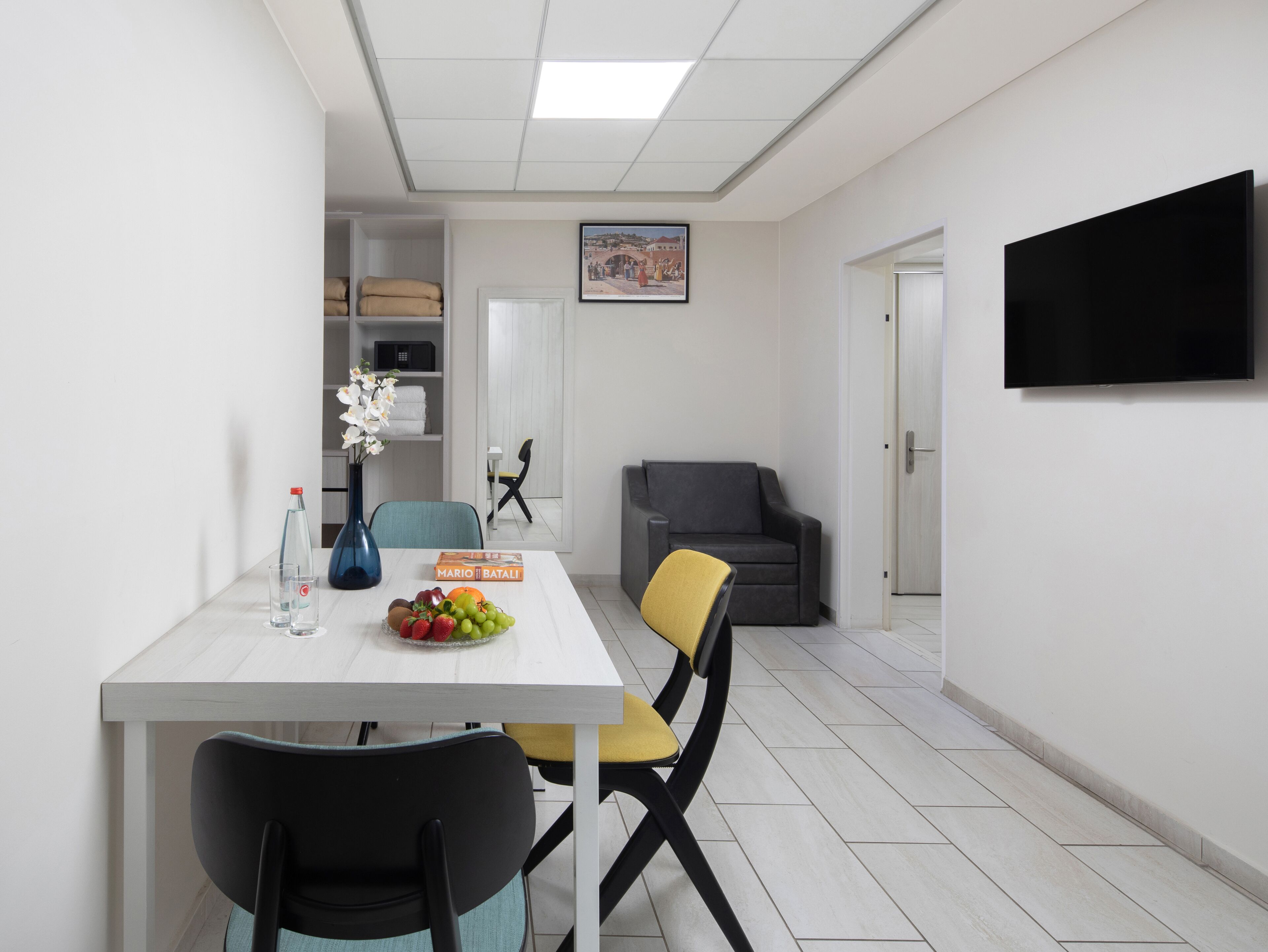 Uma sala de jantar moderna com uma mesa branca, duas cadeiras, um vaso de flores e uma cesta de frutas. Um sofá cinzento e uma televisão estão ao fundo.