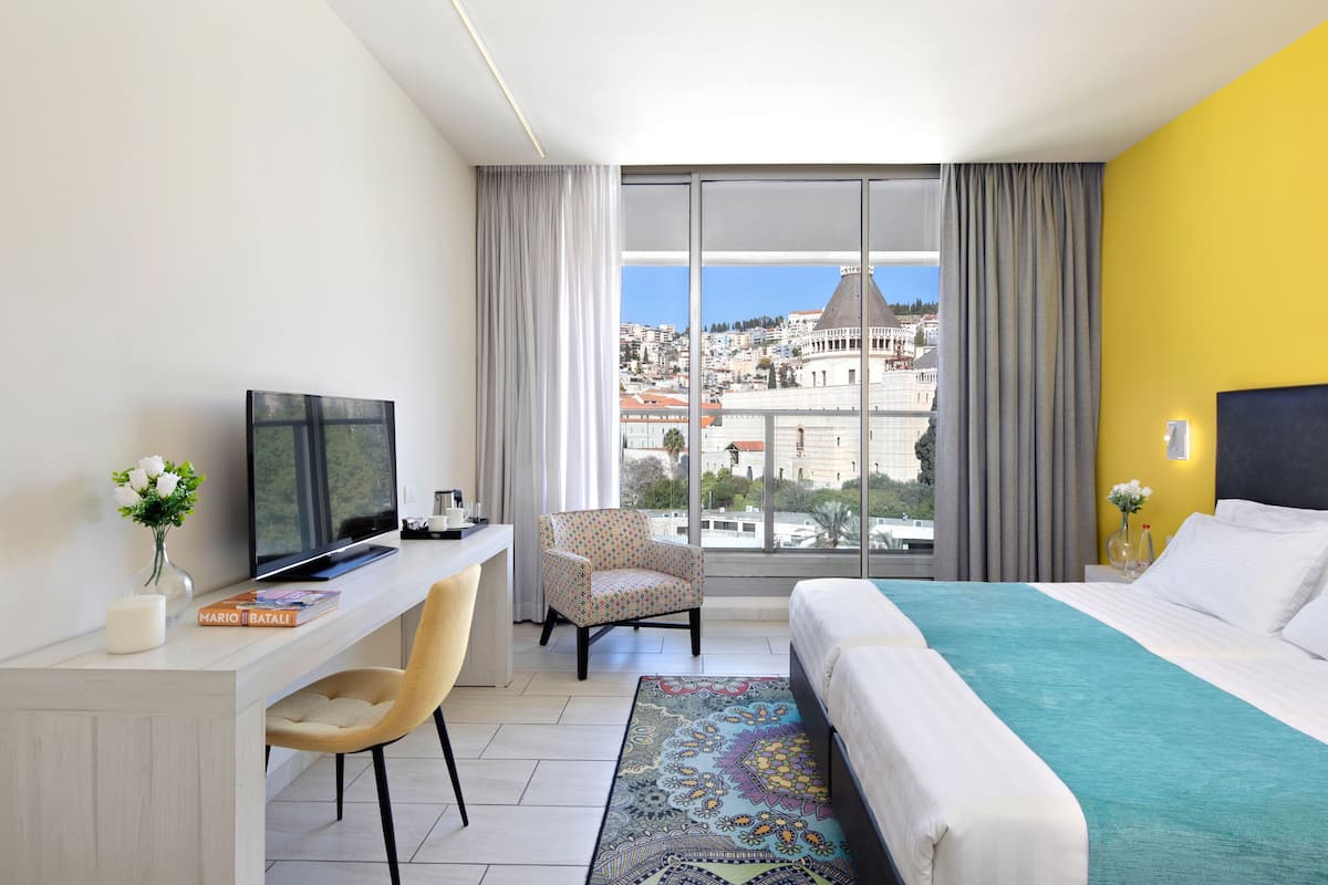 Ein Hotelzimmer mit großem Fenster, einem Bett, einem Schreibtisch mit Fernseher, einem Sessel und Blick auf die Stadt.