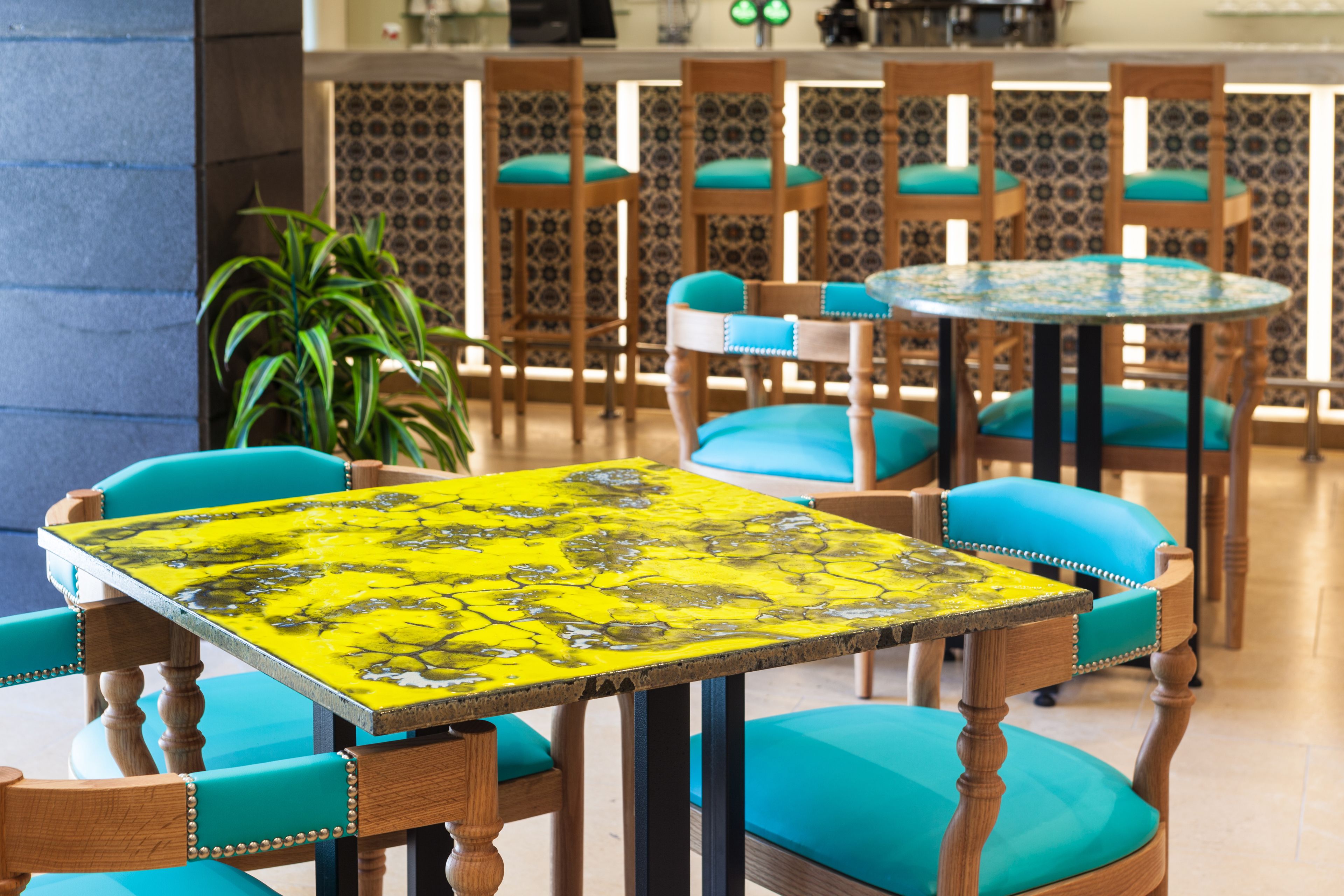 A dining area with wooden chairs and tables, a potted plant, and patterned wallpaper.