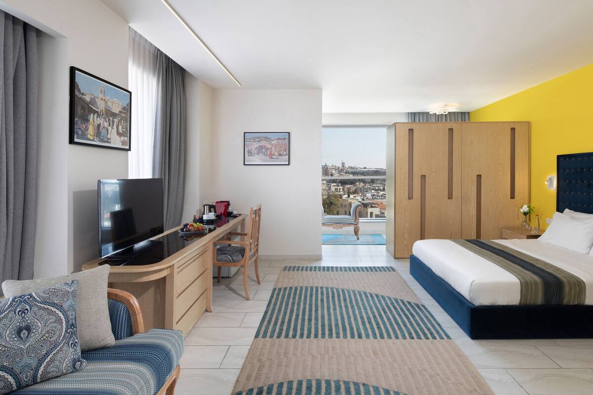 Ein modernes Hotelzimmer mit Bett, Schreibtisch, Fernseher und einem Balkon mit Blick auf die Stadt.