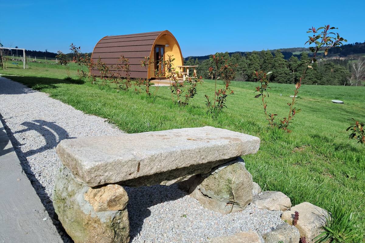 A small, single-story, semi-circular wooden structure with a porch, surrounded by a grassy field and a gravel path.