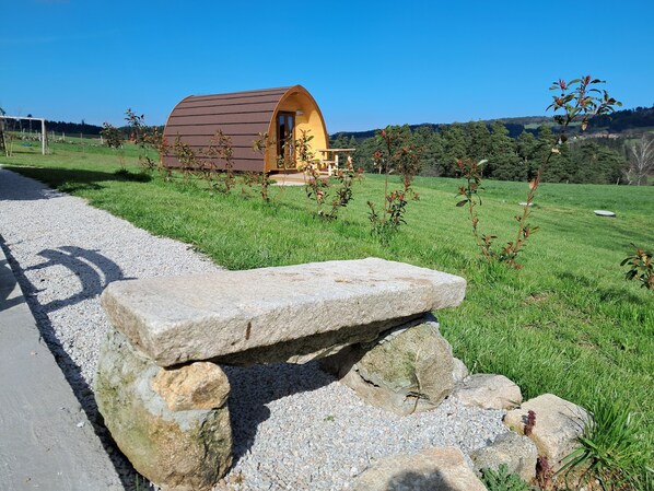 A small, single-story, semi-circular wooden structure with a porch, surrounded by a grassy field and a gravel path. - Les Roulottes de la Ferme des Chanaux (Saint-Julien-d'Ance)