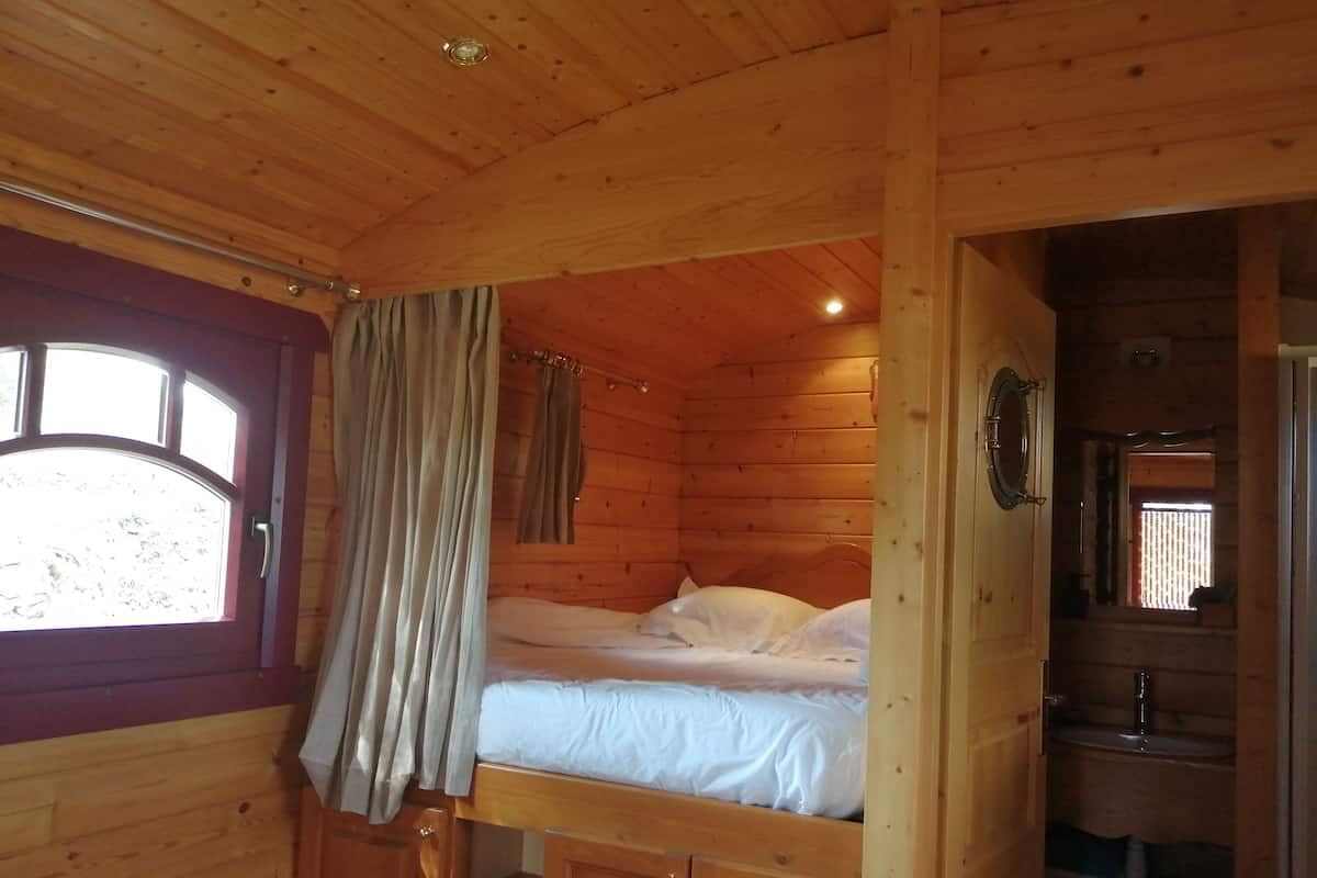 A wooden cabin with a bunk bed, a window with curtains, and a small bathroom with a sink and mirror.