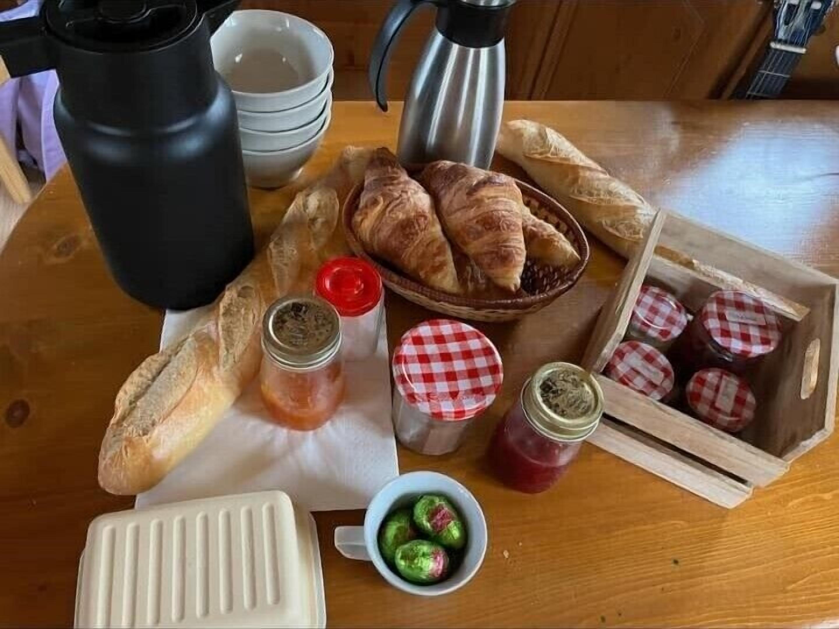 Ein Holztisch mit Croissants, Marmeladengläsern, einer Kaffeekanne und einer Gebäckauslage.