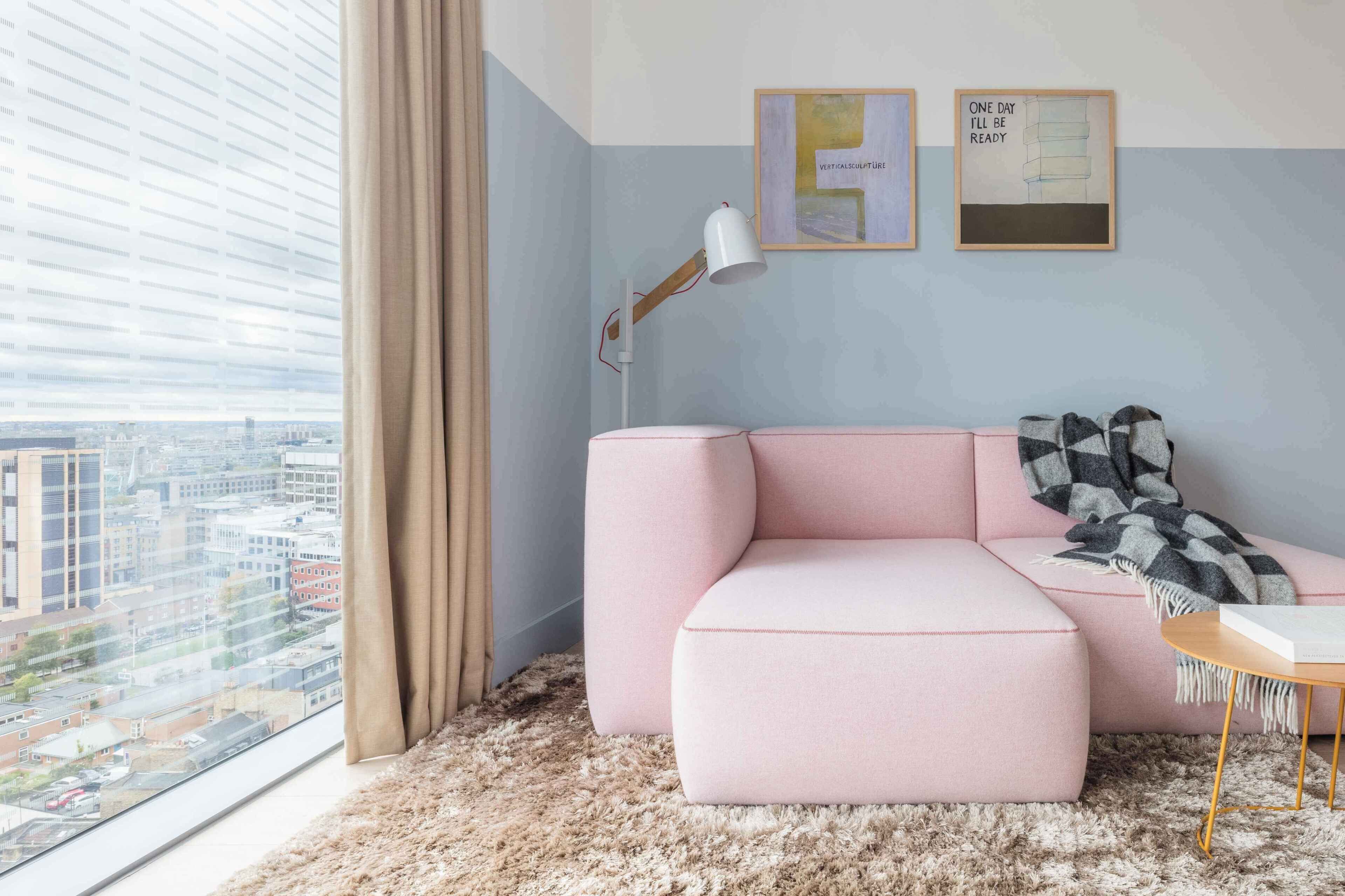 A modern living room with a pink sectional sofa, a small side table, a floor lamp, and city views through large windows.