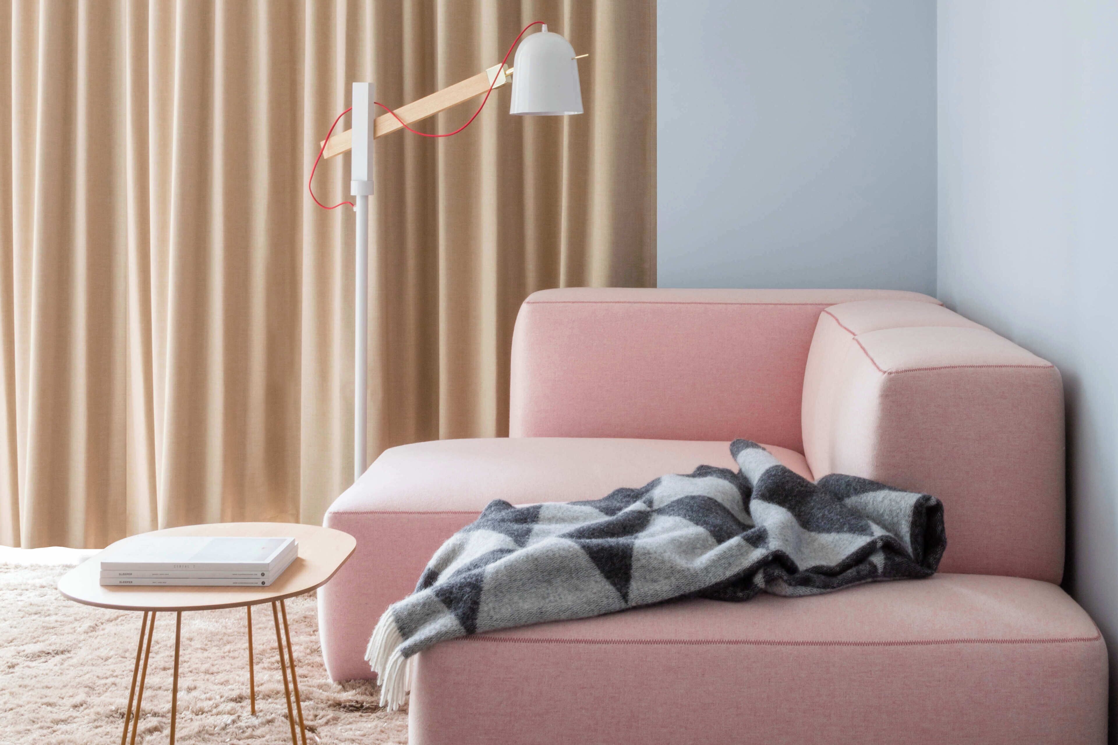 Una habitación con un sofá rosa, una pequeña mesa redonda con un libro, una lámpara y una manta.