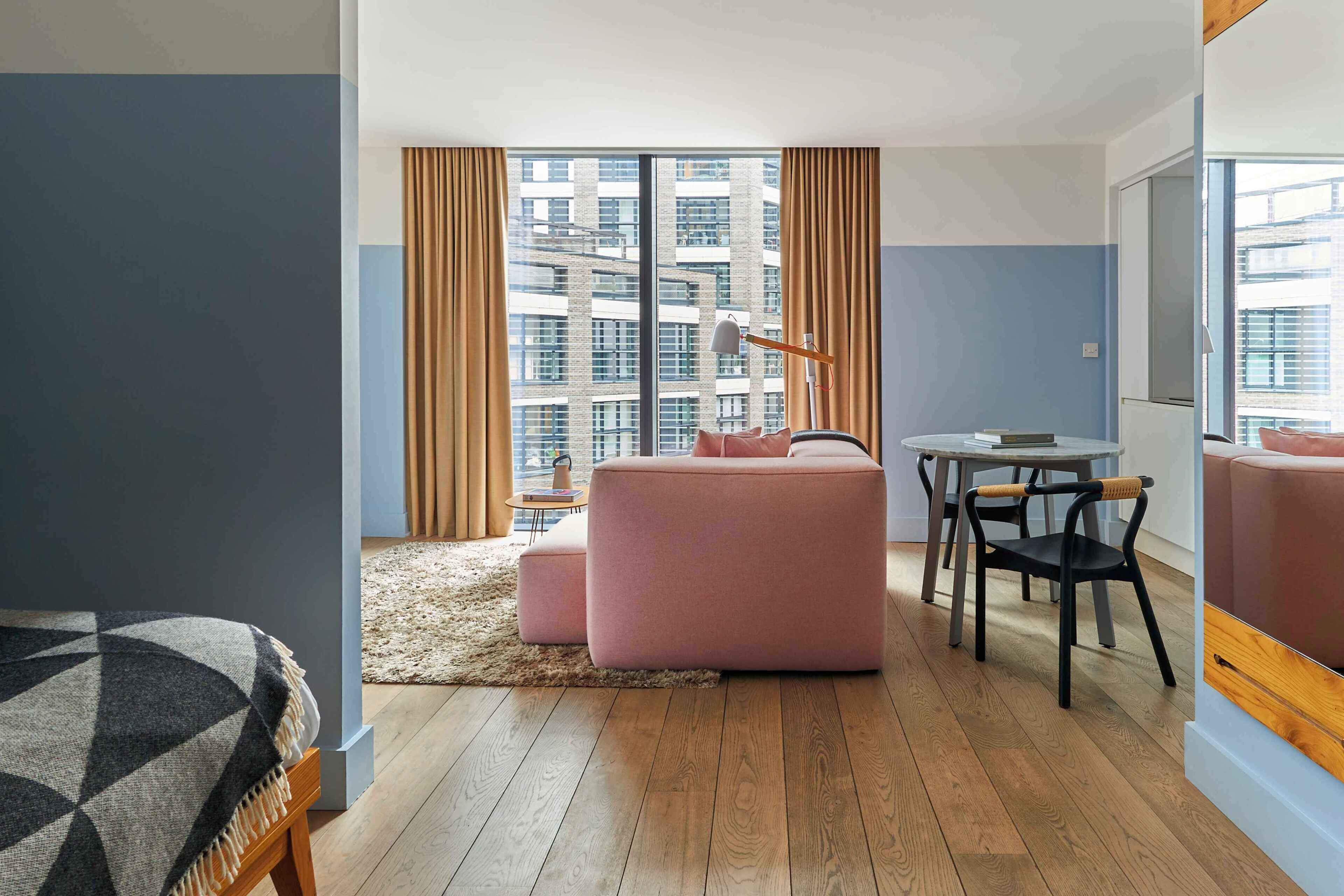Une chambre d’hôtel moderne avec une grande fenêtre, un canapé rose, une petite table avec une lampe et un plancher en bois.