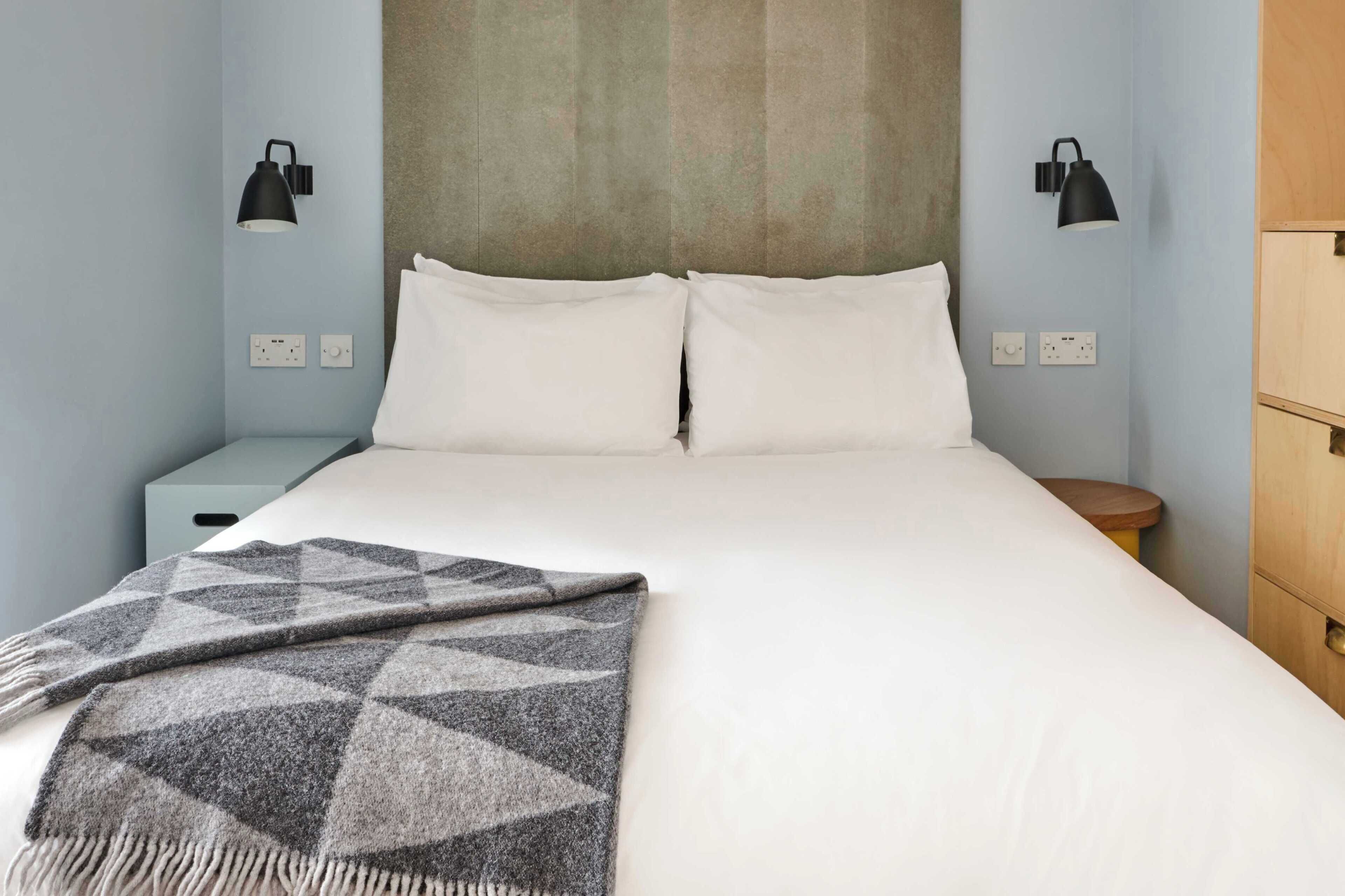 Un lit avec du linge de lit blanc, un plaid gris à motifs et deux lampes noires fixées au mur.