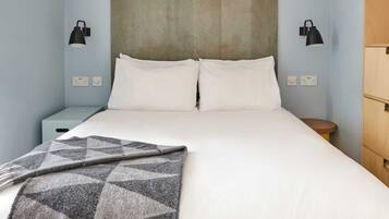 Un lit avec du linge de lit blanc, un plaid gris à motifs et deux lampes noires fixées au mur.