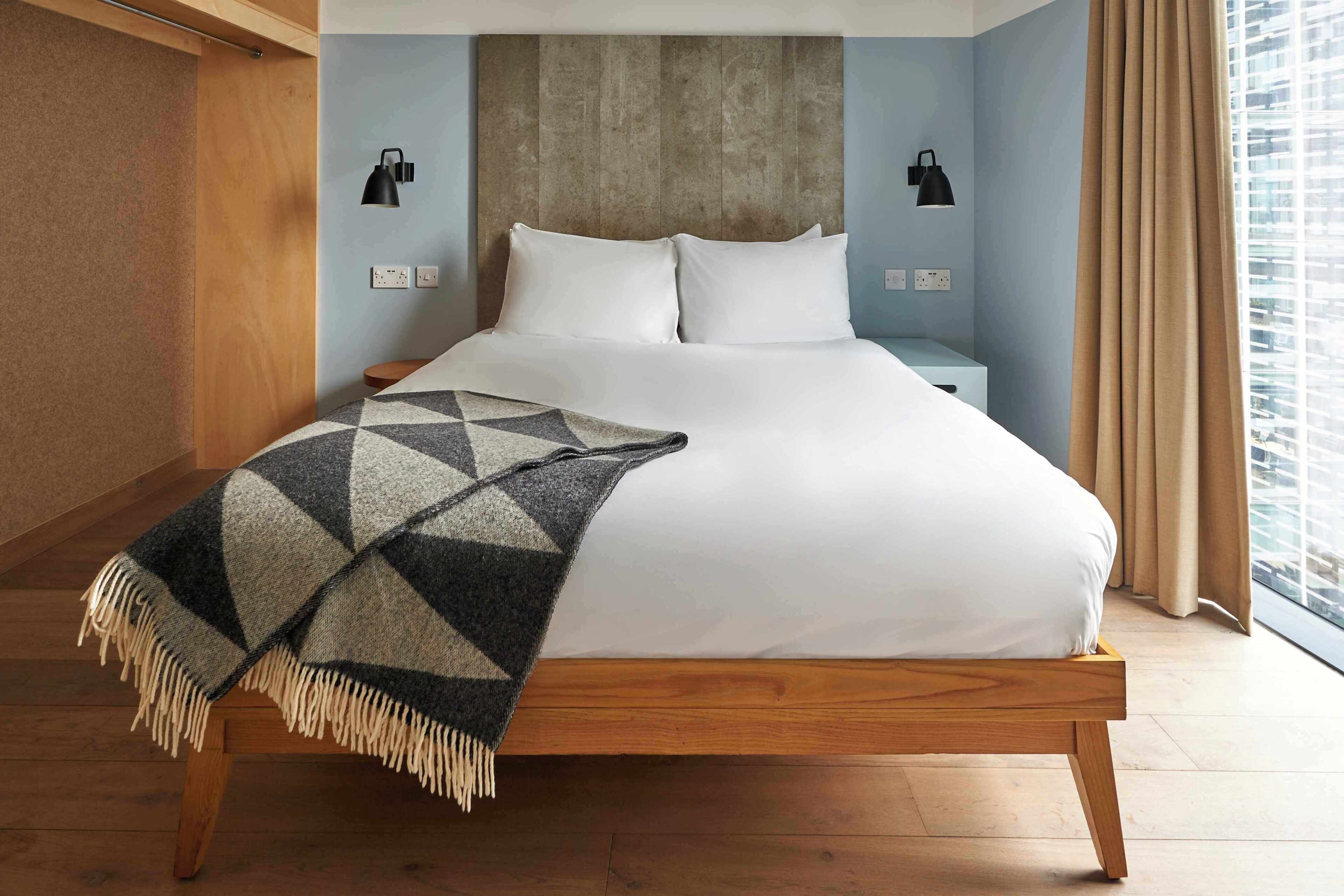 A hotel room with a bed, a wooden headboard, two bedside tables, a blanket, and a view of the city.