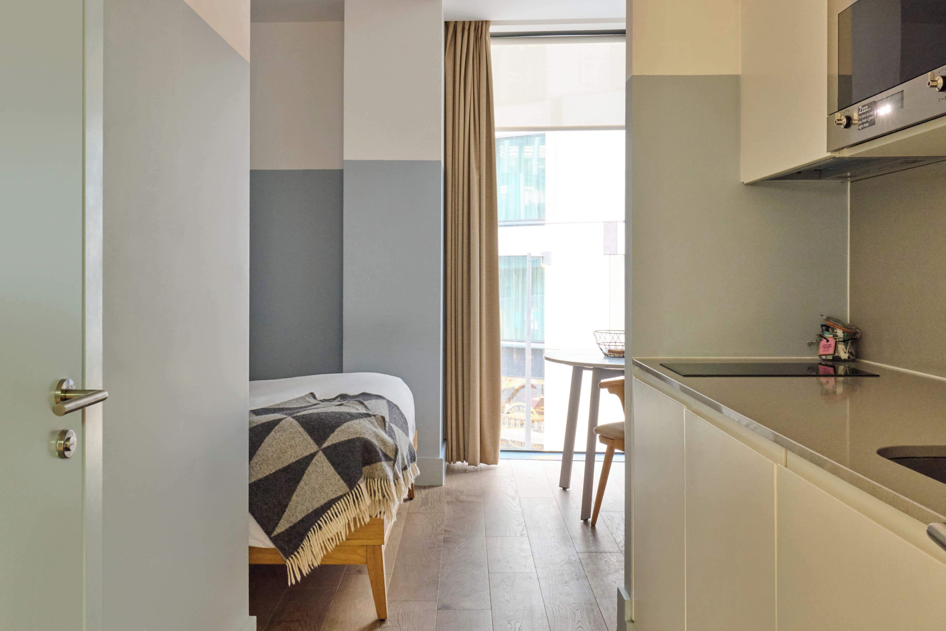 A hotel room with a bed, a dining table, a kitchenette, and a view through a window.