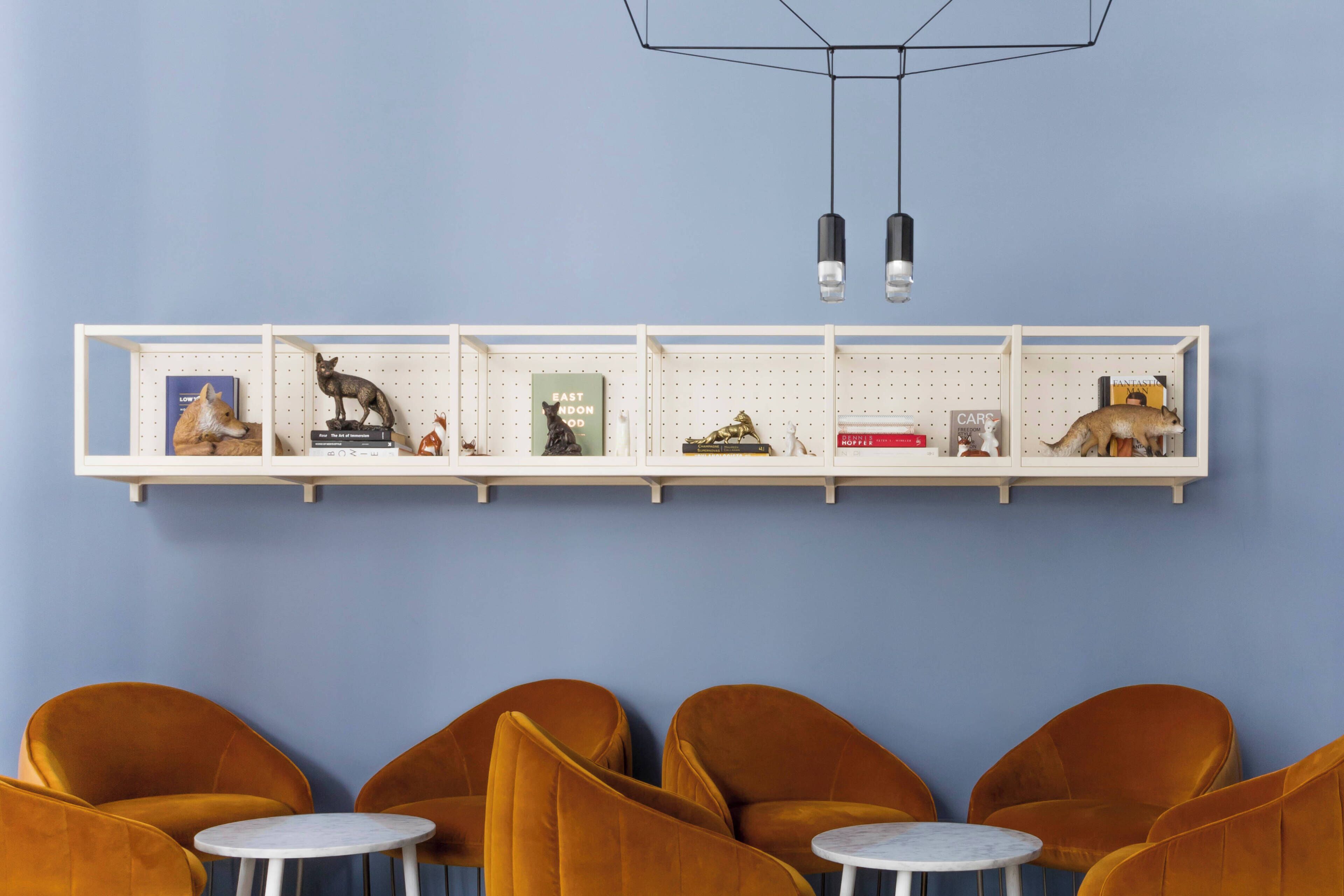 A room with a blue wall, a white shelf with various items, and four mustard-colored chairs with small round tables.