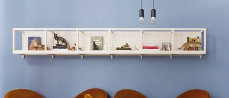 Una habitación con una pared azul, un estante blanco con diversos objetos y cuatro sillas color mostaza con pequeñas mesas redondas.