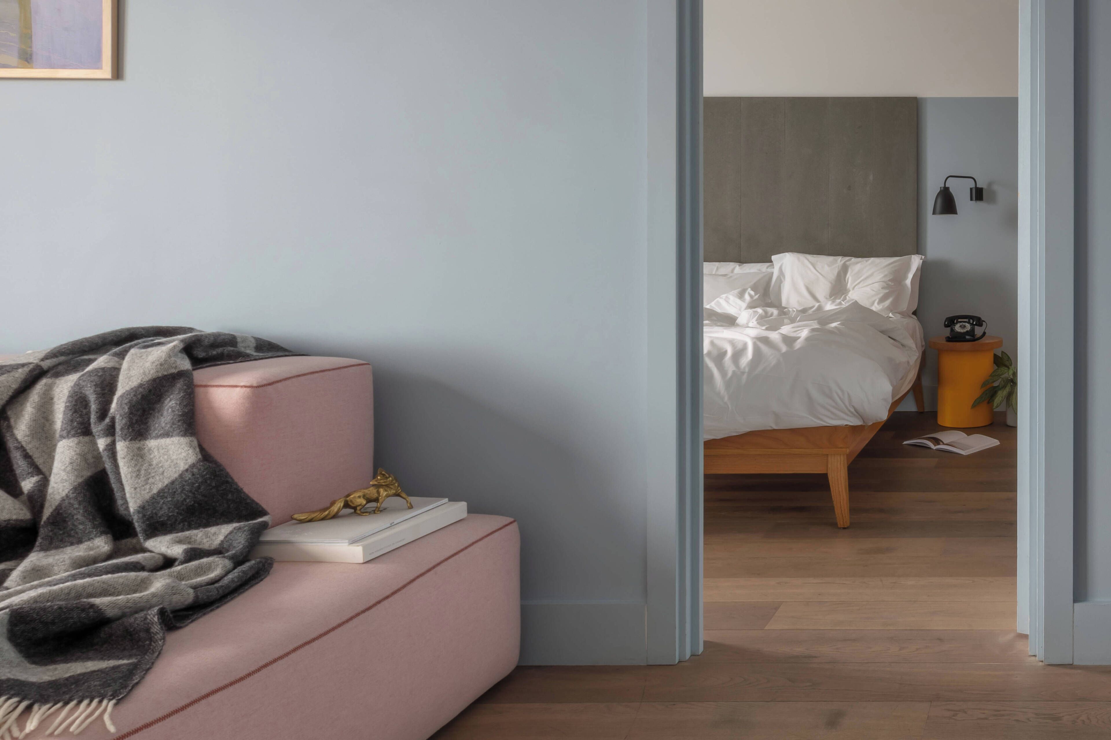 Una habitación con un sofá rosa, una manta gris, una mesita con un libro y una estatua dorada, y una cama de madera con ropa de cama blanca.