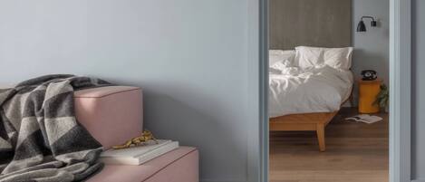 Una habitación con un sofá rosa, una manta gris, una mesita con un libro y una estatua dorada, y una cama de madera con ropa de cama blanca.