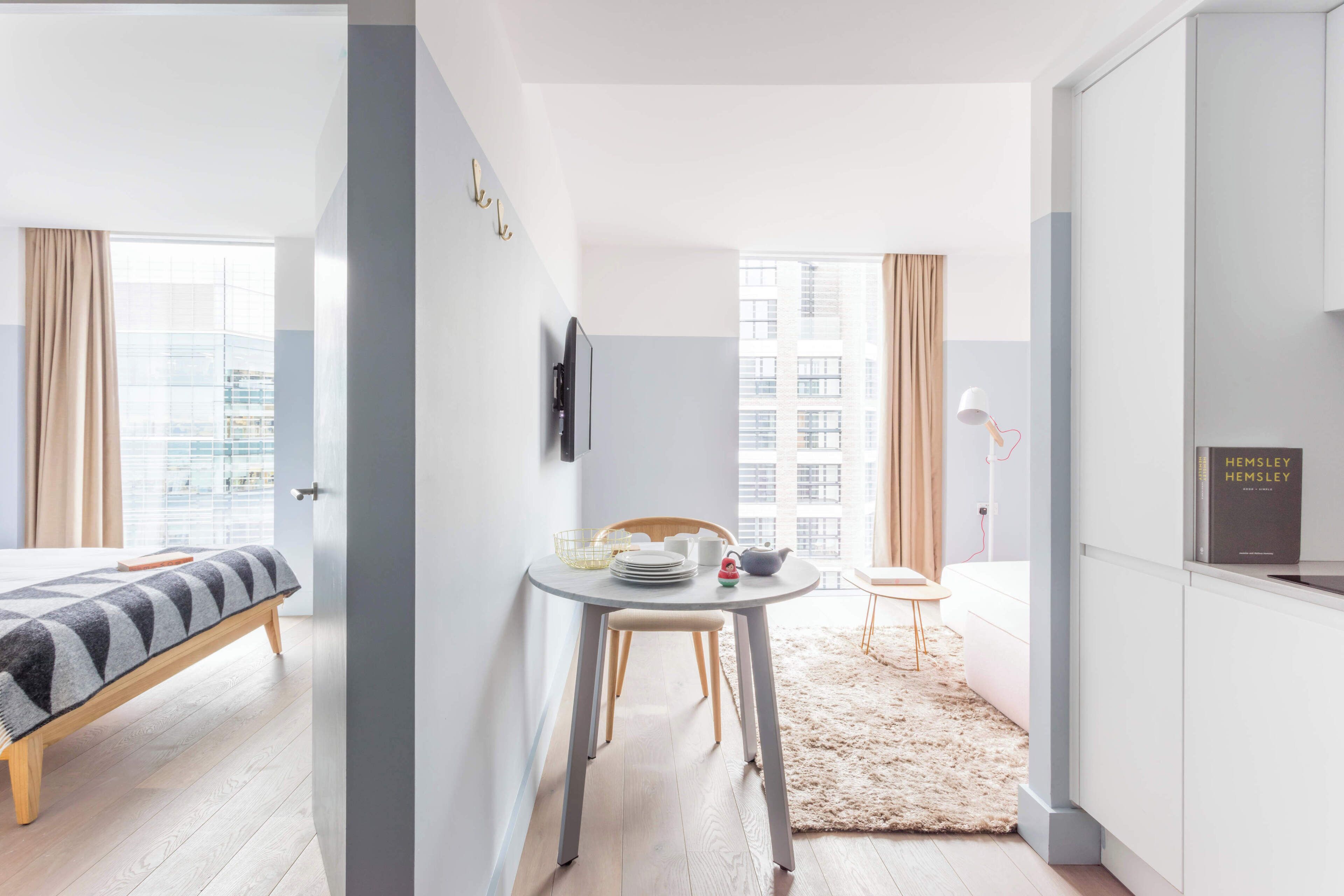 Een moderne hotelkamer met een bed, een zithoek met tafel en stoelen, een klein keukenhoekje en grote ramen met uitzicht op de stad.