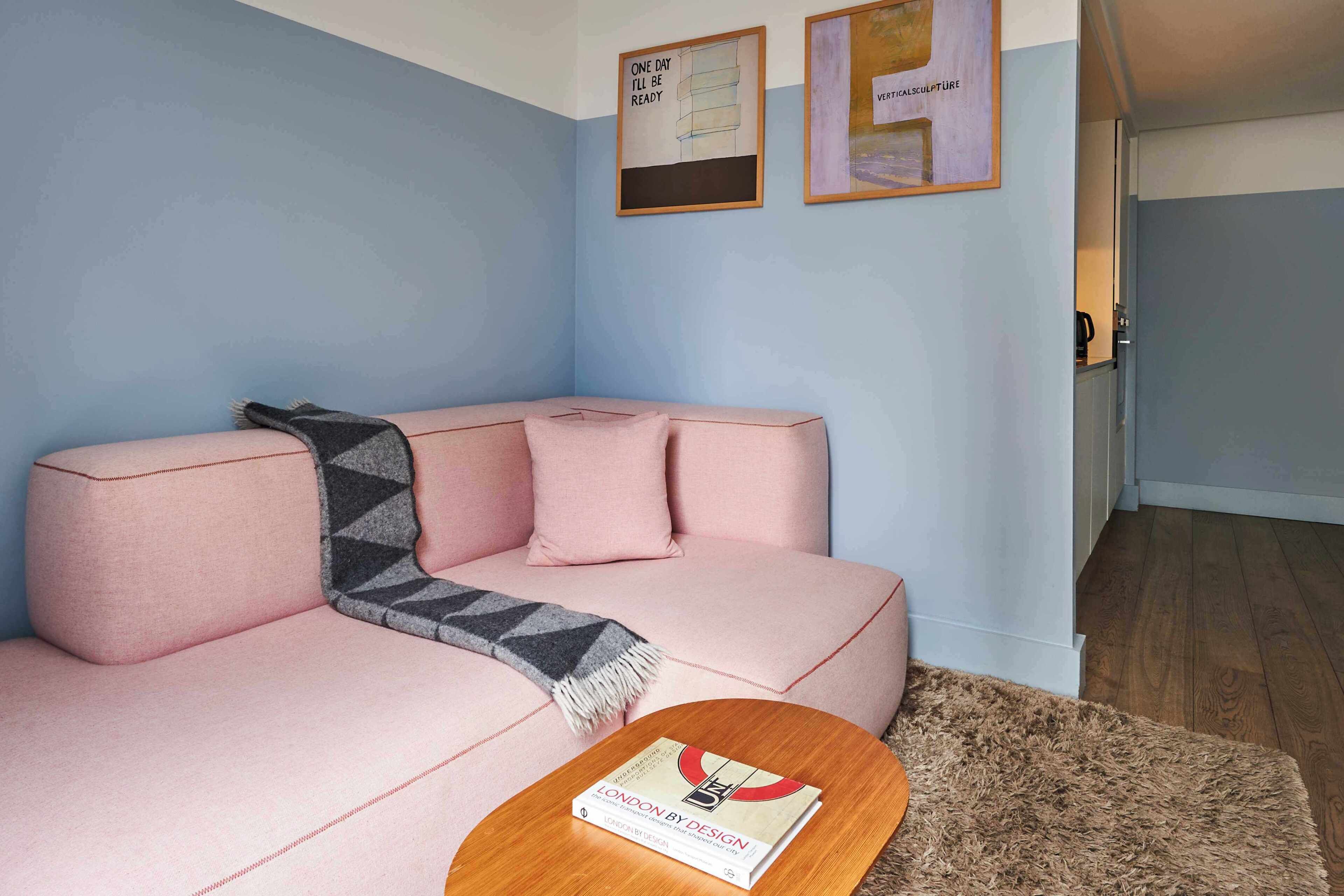 A pink sofa with a patterned throw, a wooden coffee table with a book, and a kitchen visible in the background.