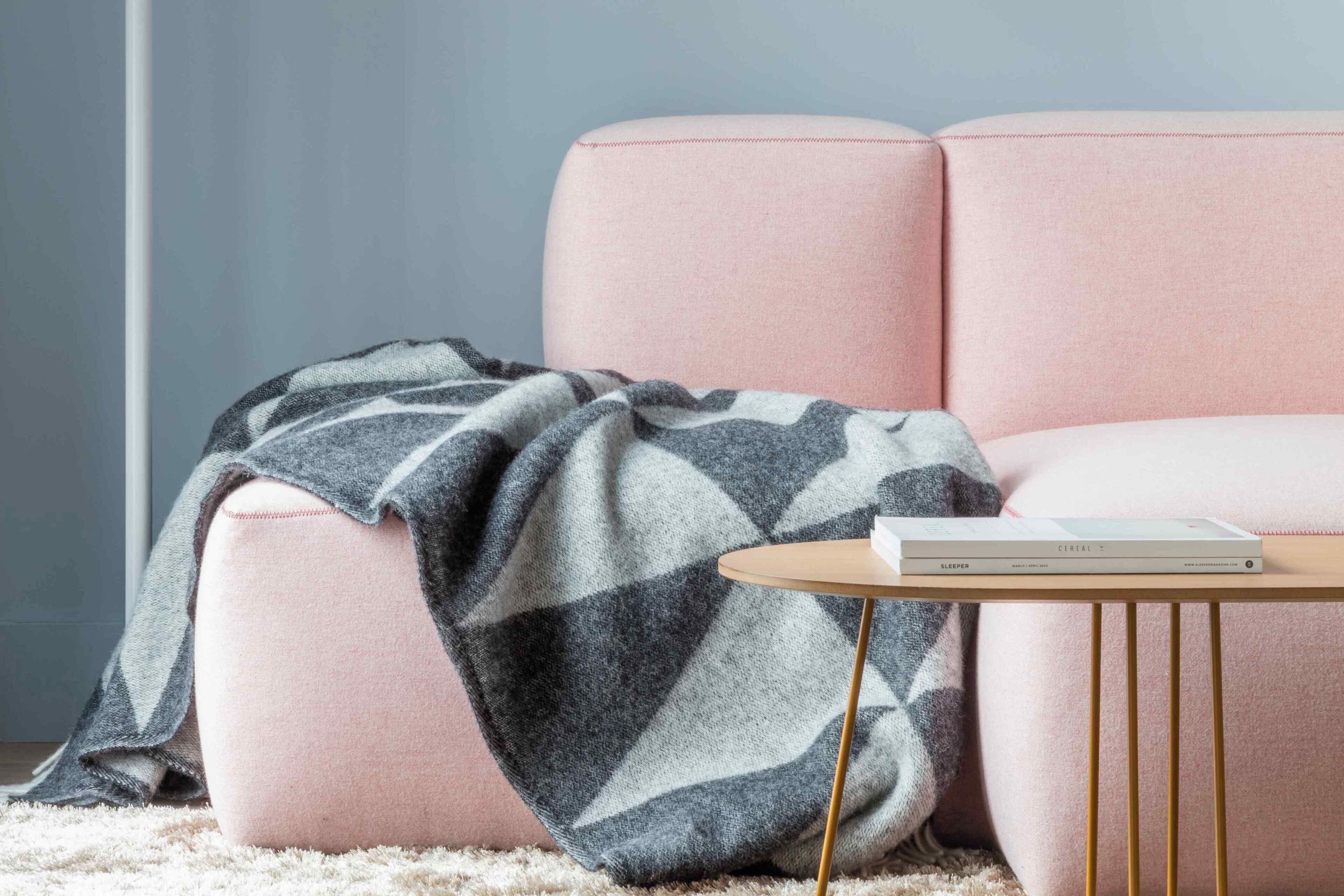 Un sofá rosa con una manta, una mesita redonda con un libro y una alfombra de color claro.